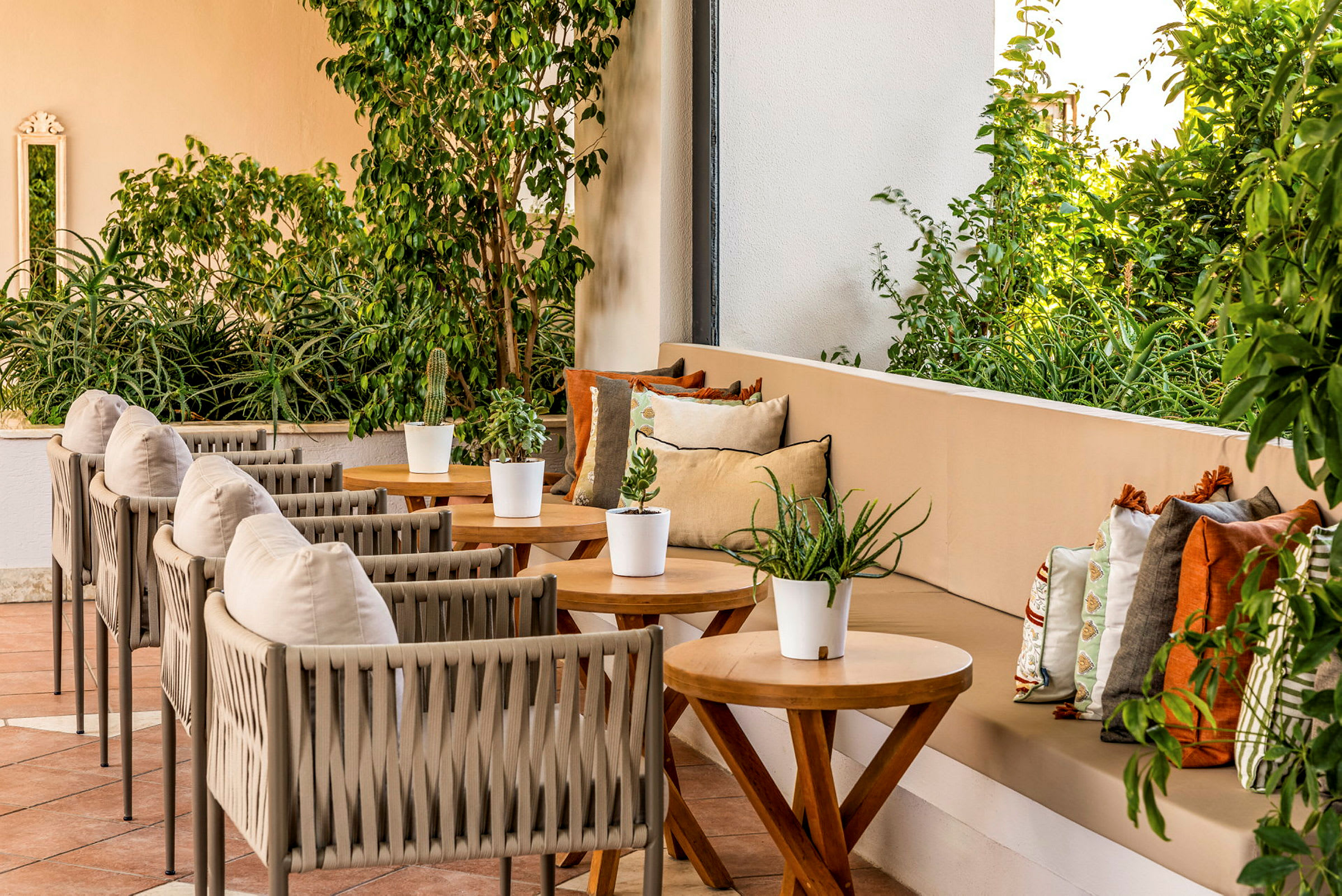 En terrasse med flere borde og stole samt en sofa med farverige puder. Planter og grønt omkranser området. (AI-genereret tekst)