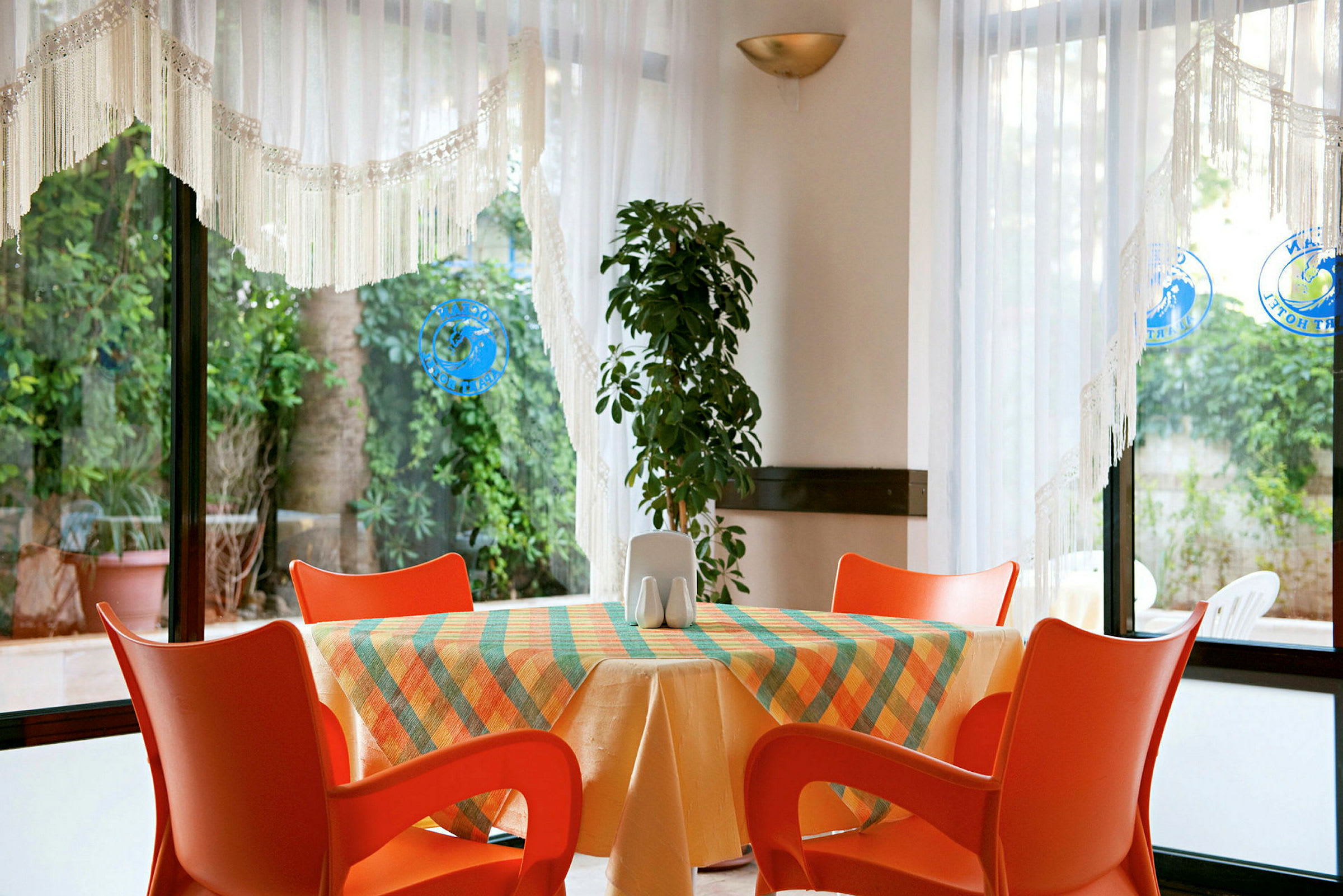 Et bord med farverig dug og orange stole foran et vindue med udsigt til grønne planter. (AI-genereret tekst)