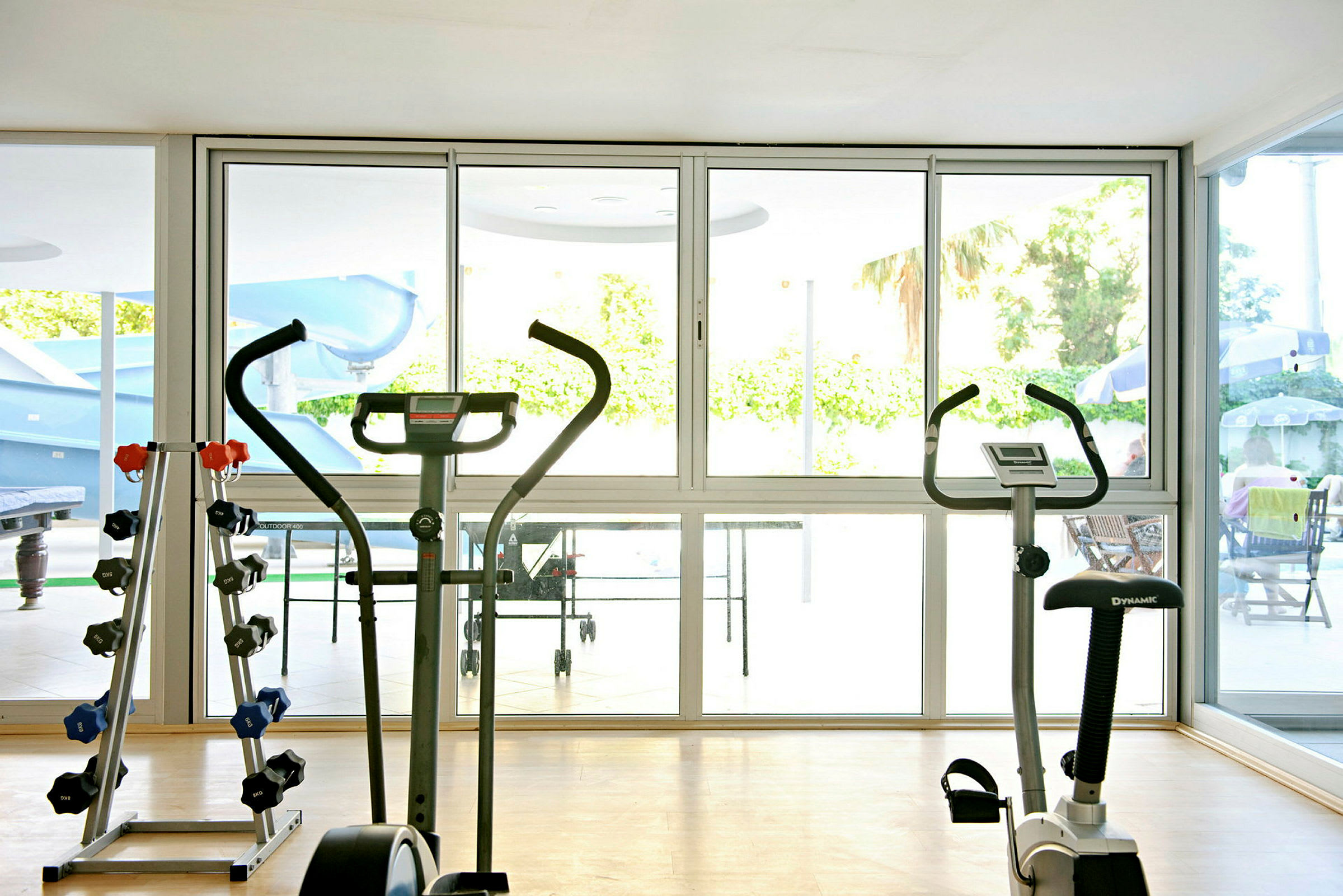 Et fitnesscenter med motionscykler og håndvægte, udsigt til et poolområde gennem glasvægge. (AI-genereret tekst)