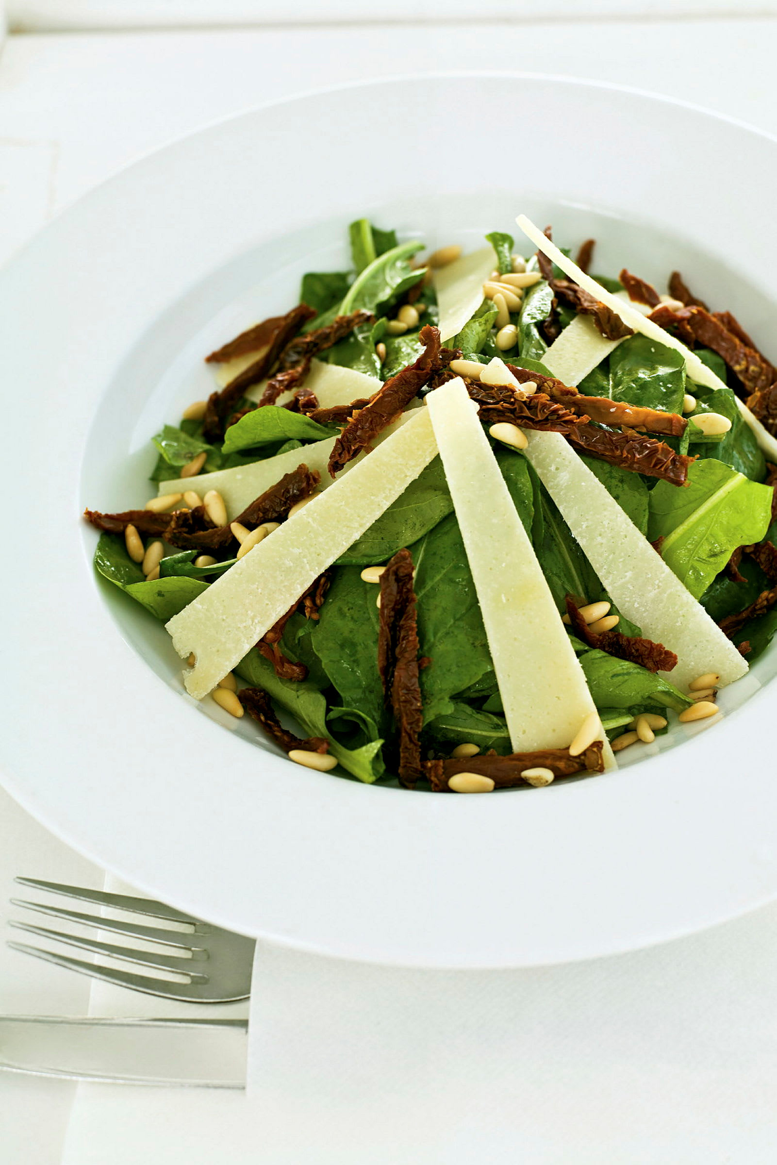 En tallerken med salat bestående af grønne blade, soltørrede tomater, pinjekerner og tynde skiver ost. (AI-genereret tekst)