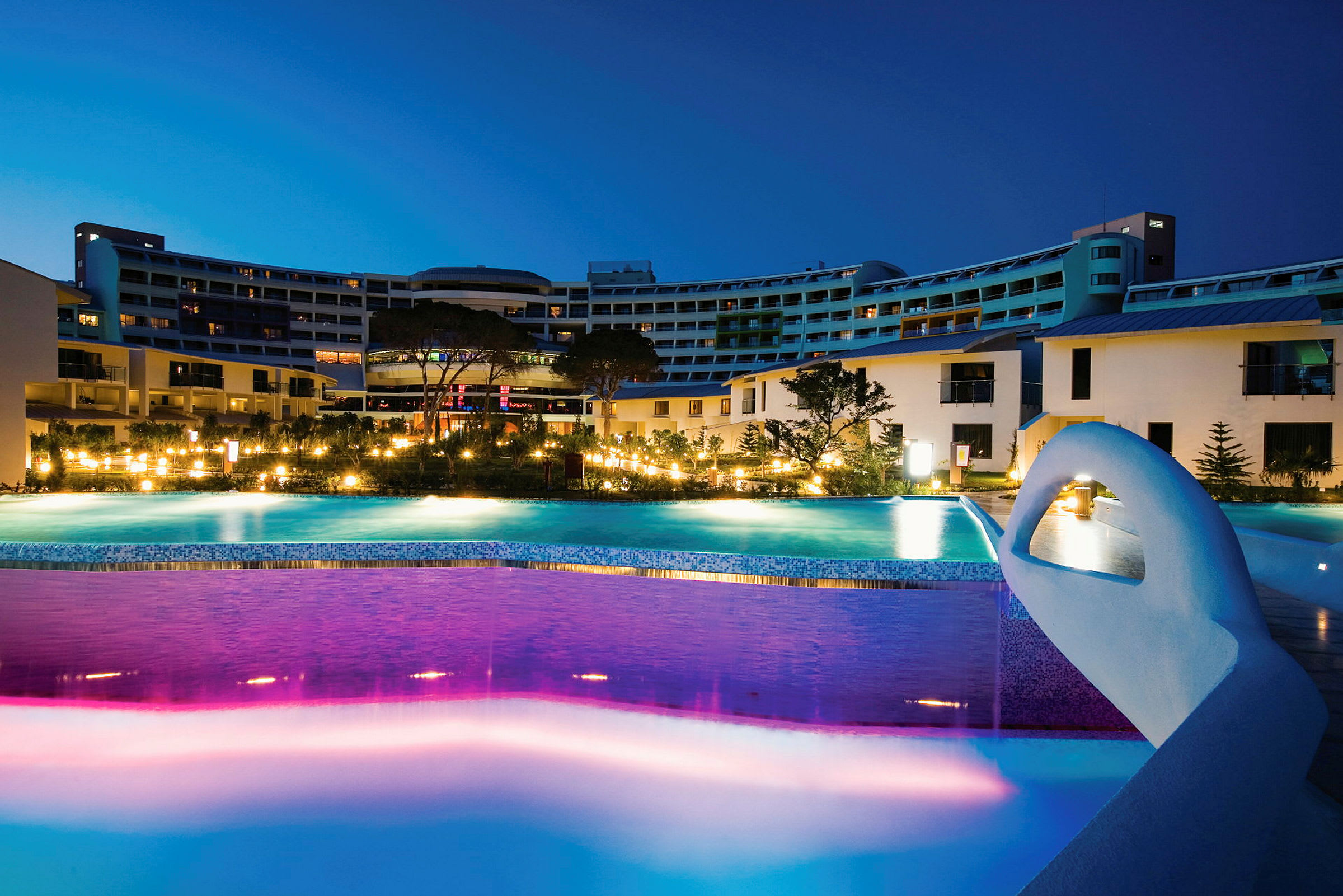 Hotelkompleks med oplyste pools om aftenen. (AI-genereret tekst)