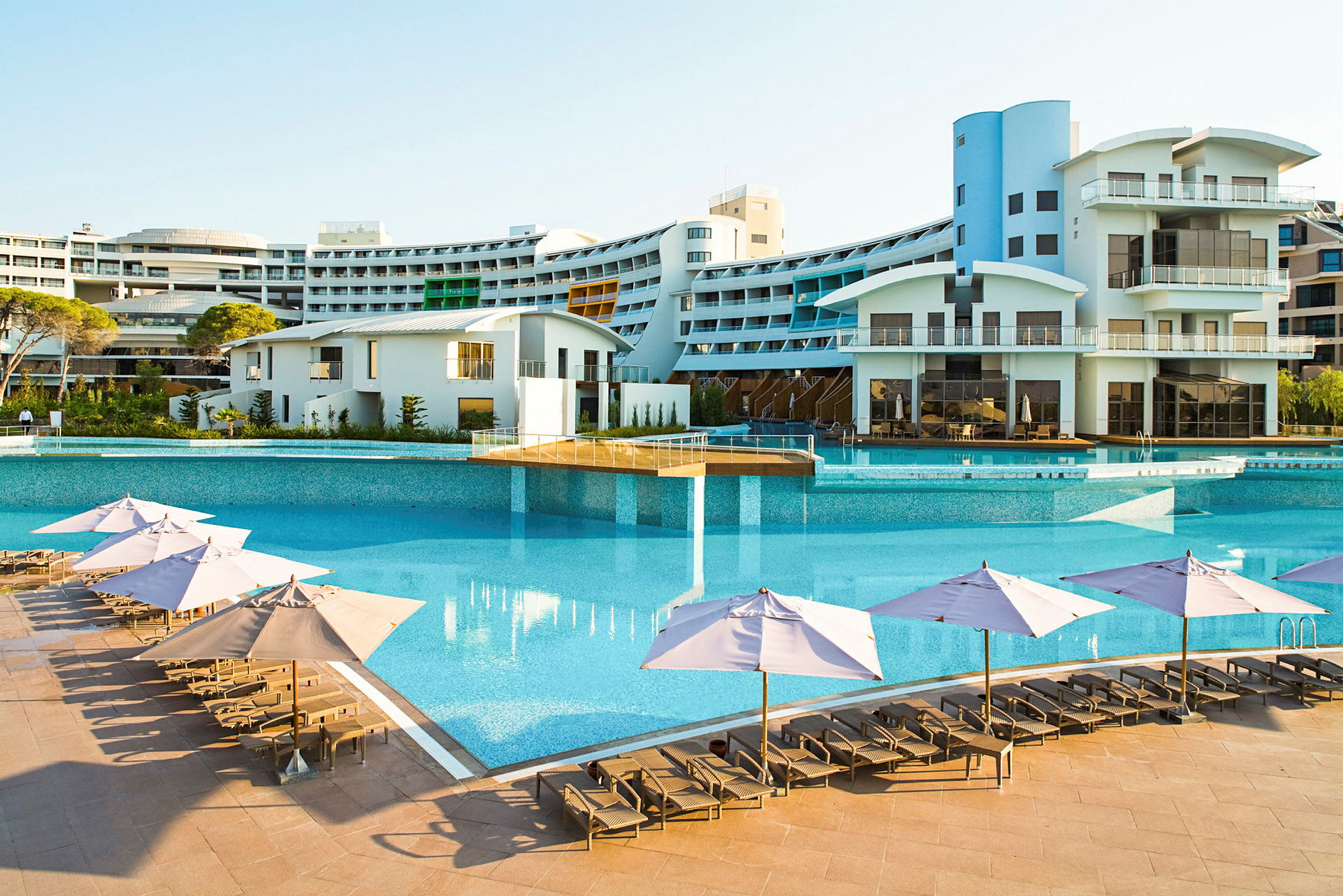 En stor udendørs pool med liggestole og parasoller foran et moderne hotel. (AI-genereret tekst)