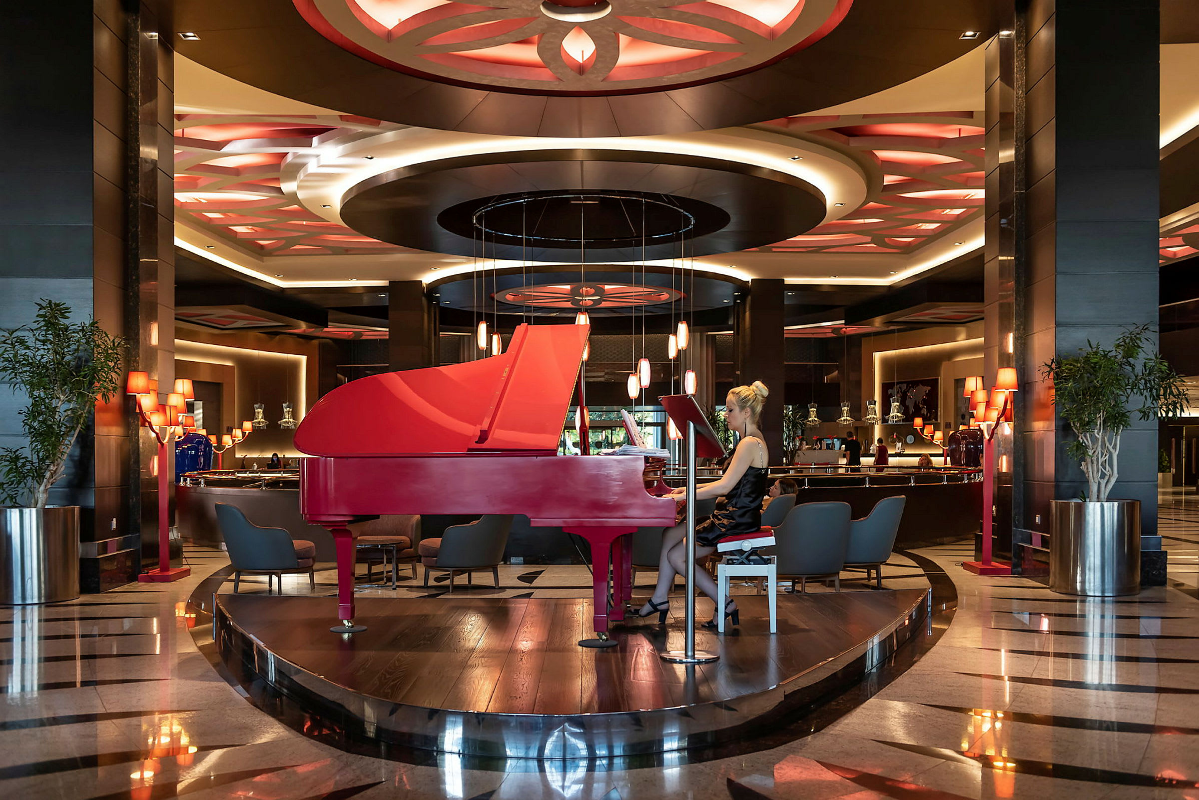 Et rødt flygel står centralt placeret i en elegant hotellobby med dekorative loftlamper. (AI-genereret tekst)