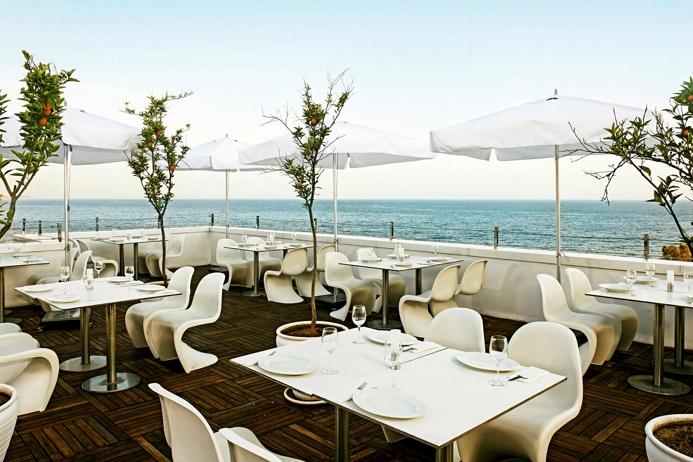 Restaurant med udendørsservering, hvide borde og stole samt hvide parasoller. Baggrund med udsigt over havet. (AI-genereret tekst)