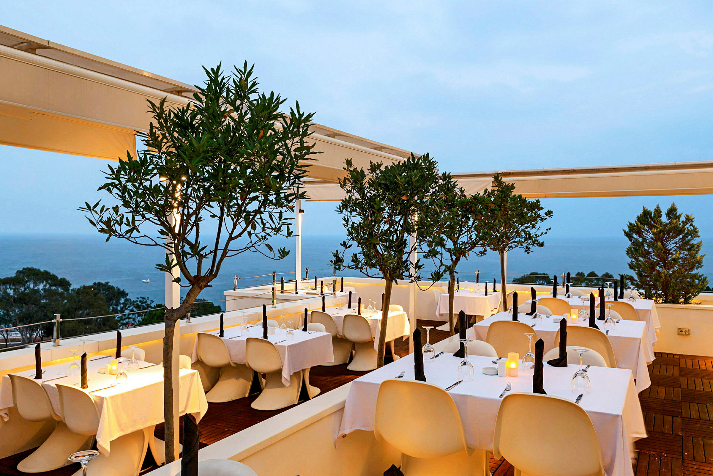 En elegant udendørsrestaurant ved havet med hvide duge på bordene og små træer som dekoration. (AI-genereret tekst)