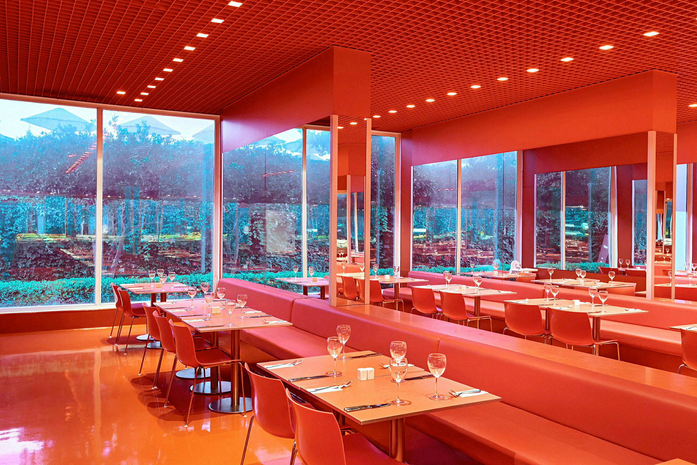En moderne restaurant med røde møbler og store glasvinduer, der vender ud mod en frodig have. (AI-genereret tekst)