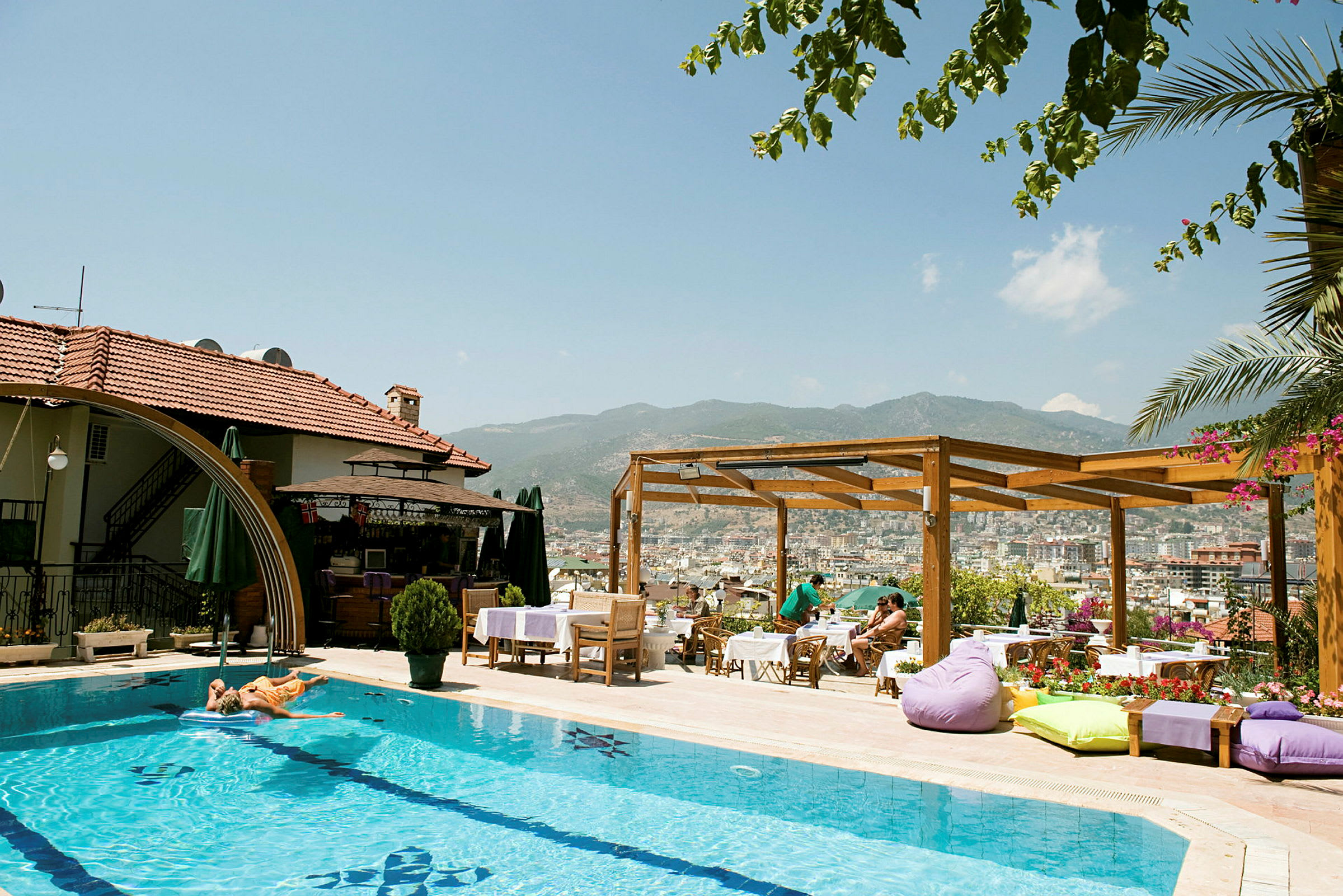 En udendørs pool ved et hotel med solsenge og parasoller. En træpergola og udsigt over en by og bjerge i baggrunden. (AI-genereret tekst)