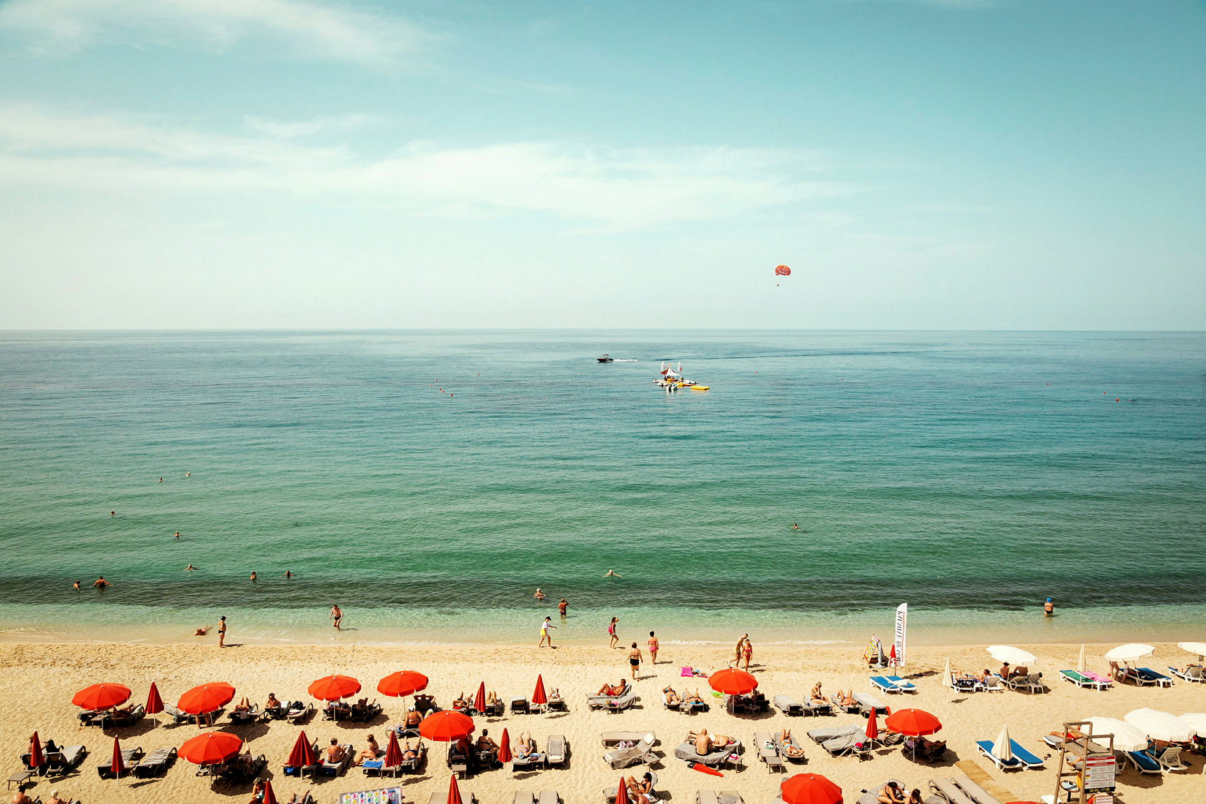 En strandscene med røde parasoller, liggestole og mennesker ved vandet. (AI-genereret tekst)