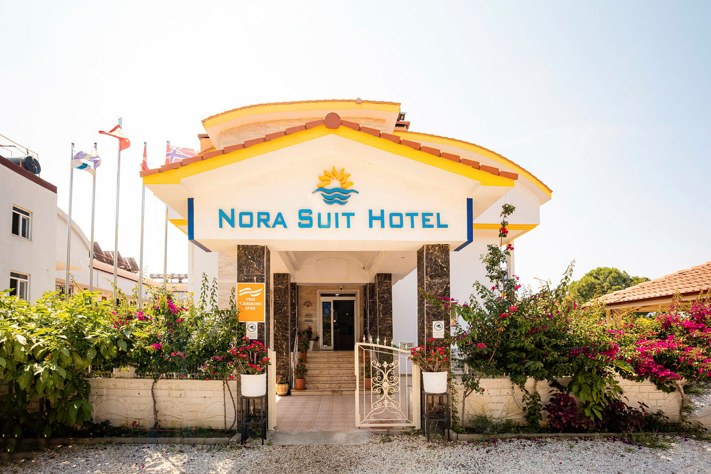 Indgangen til Nora Suit Hotel med blomster og flag i baggrunden. (AI-genereret tekst)