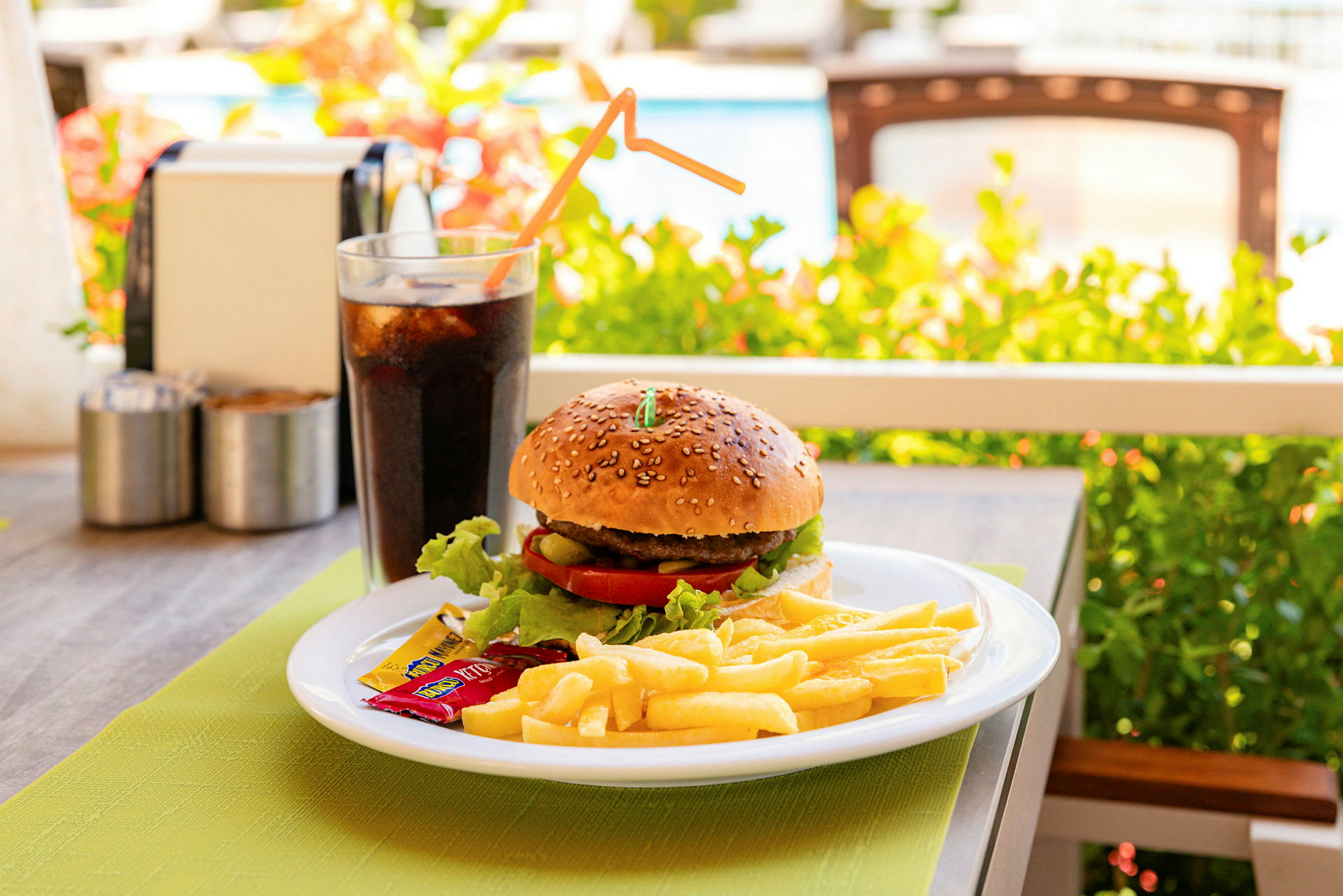En tallerken med en burger, pommes frites og krydderposer samt et glas sodavand står på et bord udenfor. (AI-genereret tekst)
