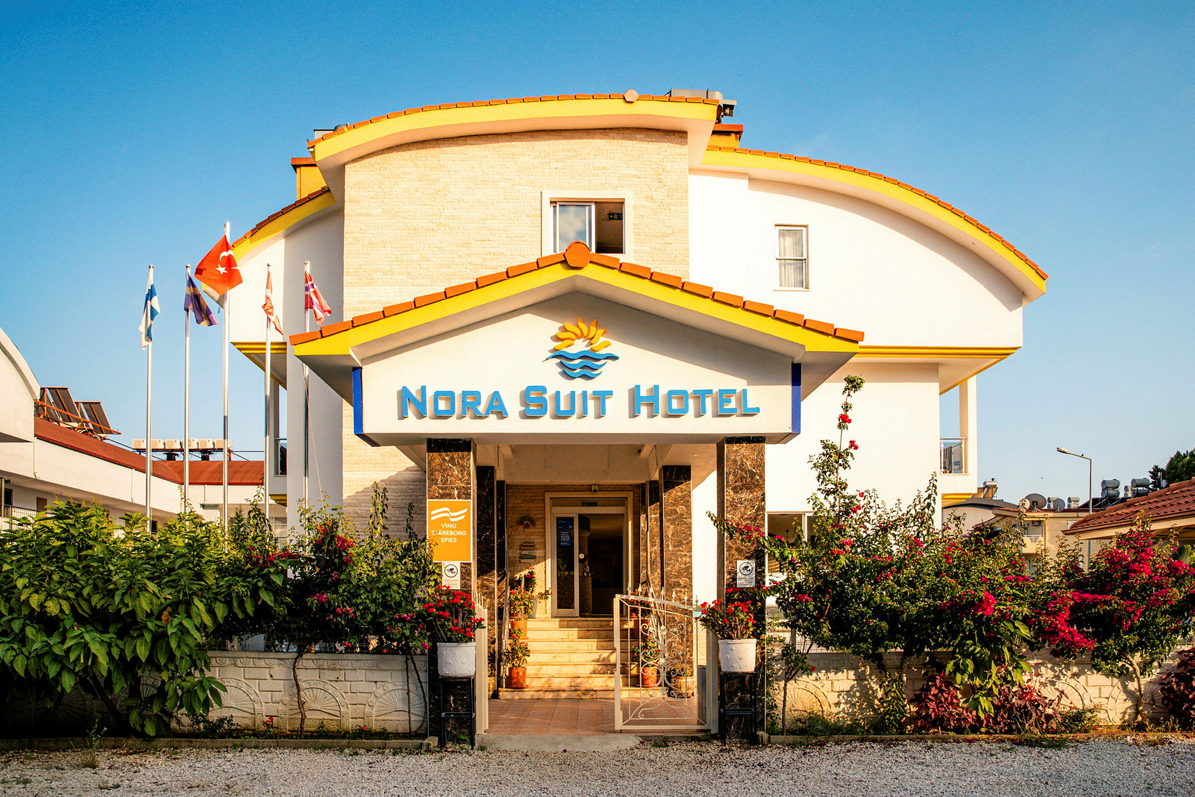 Bygningen til Nora Suit Hotel med en indgang omkranset af blomster og flag. (AI-genereret tekst)