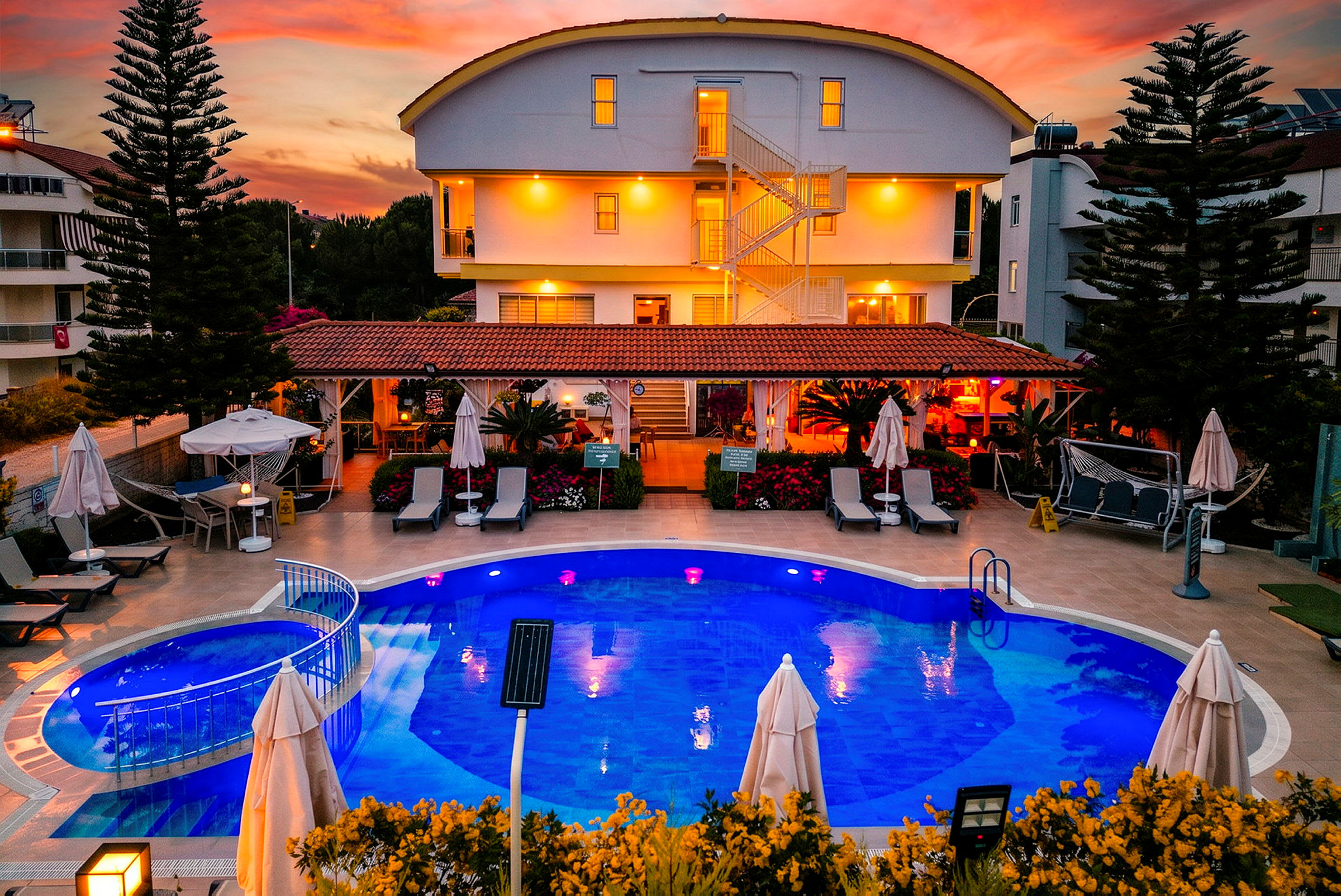 En hotelbygning med oplyst facade og en pool foran, omgivet af liggestole og parasoller under en farverig solnedgang. (AI-genereret tekst)