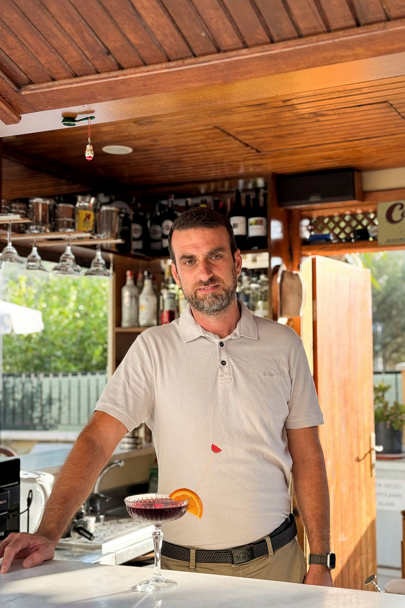En hvid skjorte ved en bar med hylder fyldt med flasker i baggrunden, samt et cocktailglas med en farvet drik foran sig. (AI-genereret tekst)