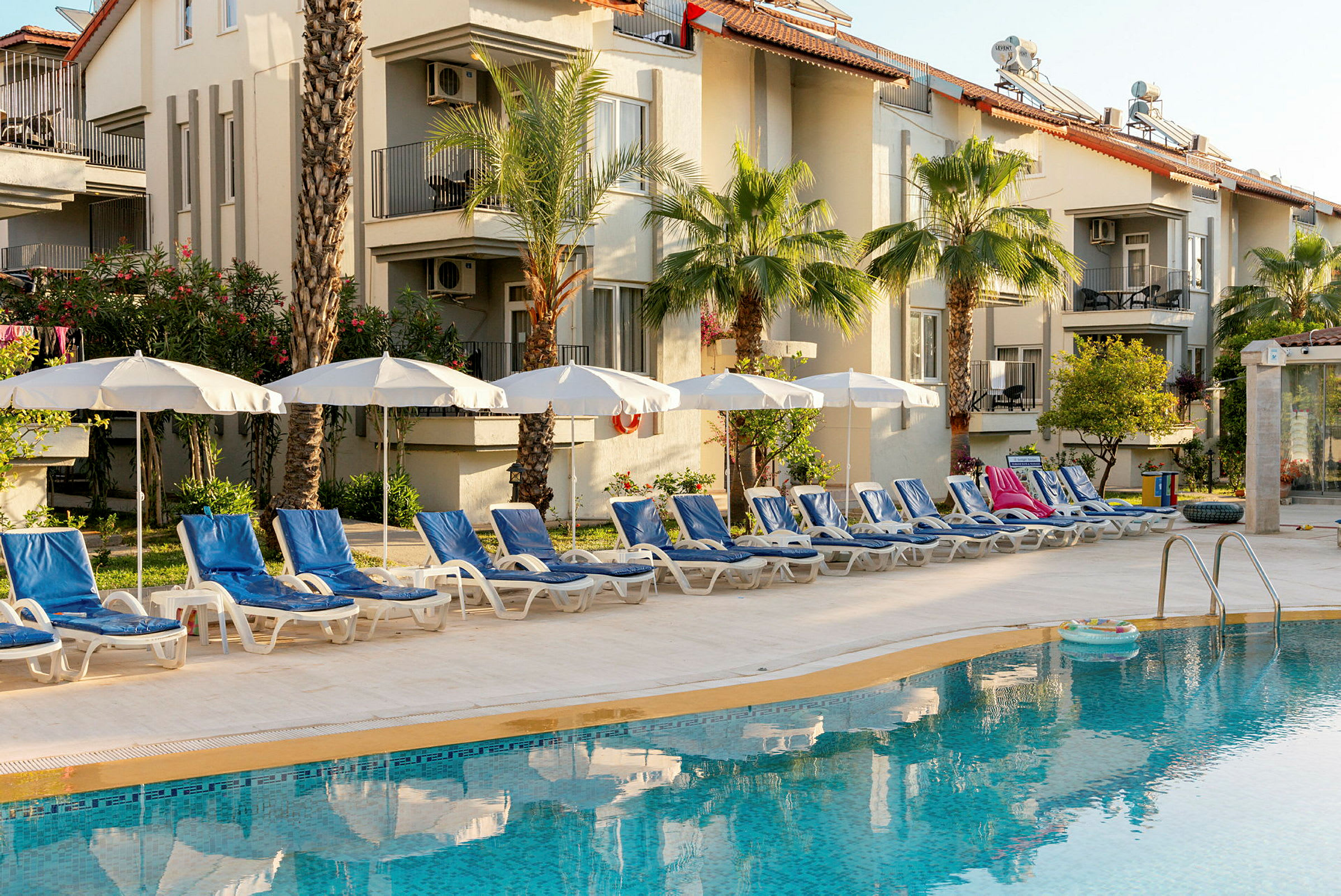 Billede af en pool med liggestole og parasoller foran et hotel. (AI-genereret tekst)