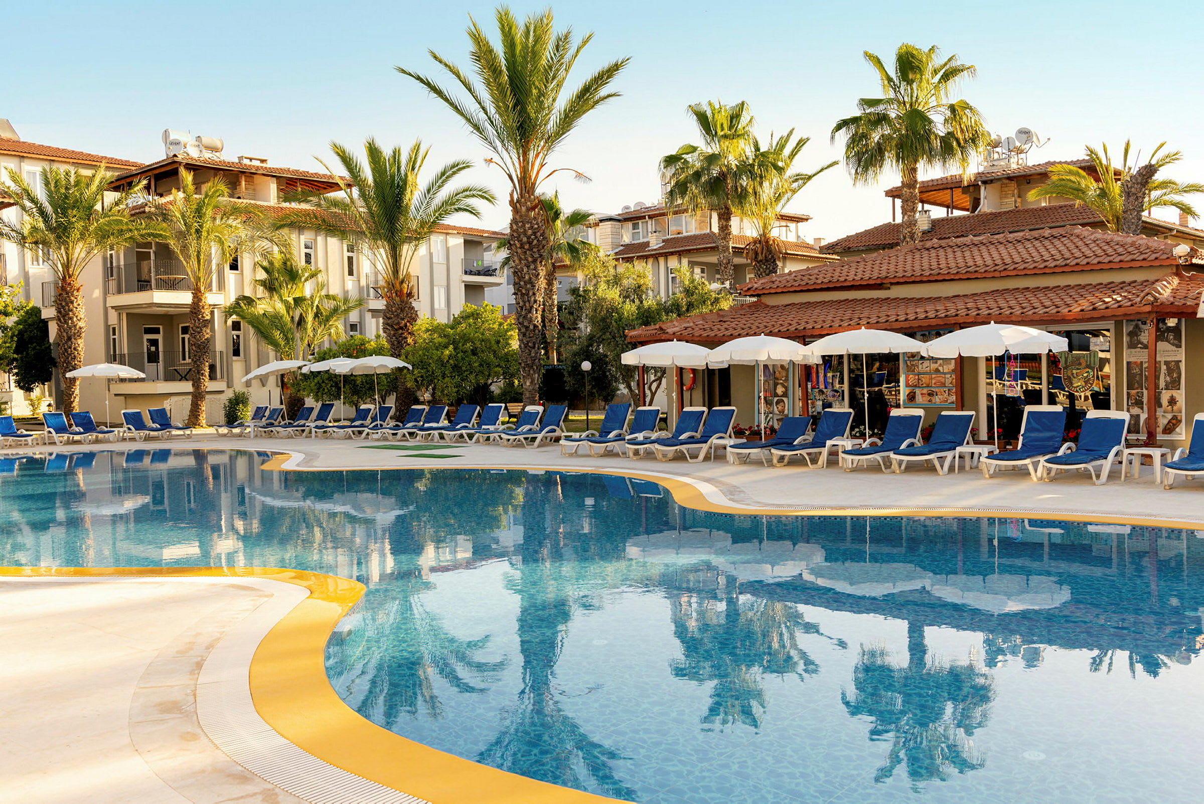 En udendørs pool med liggestole og parasoller, omgivet af palmer og hotelbygninger. (AI-genereret tekst)