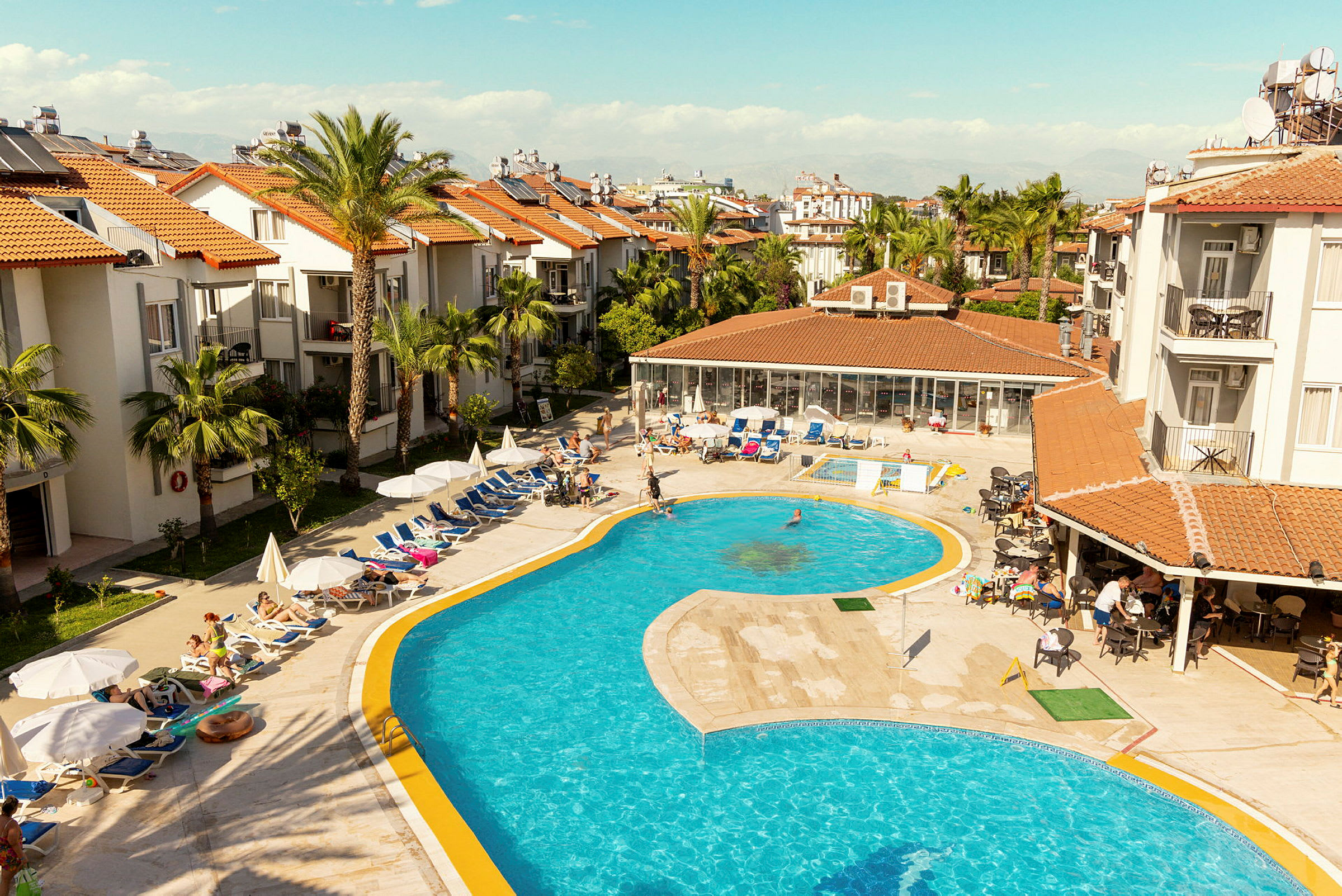 Hotelområde med pool og liggestole omgivet af bygninger med røde tegltage. (AI-genereret tekst)
