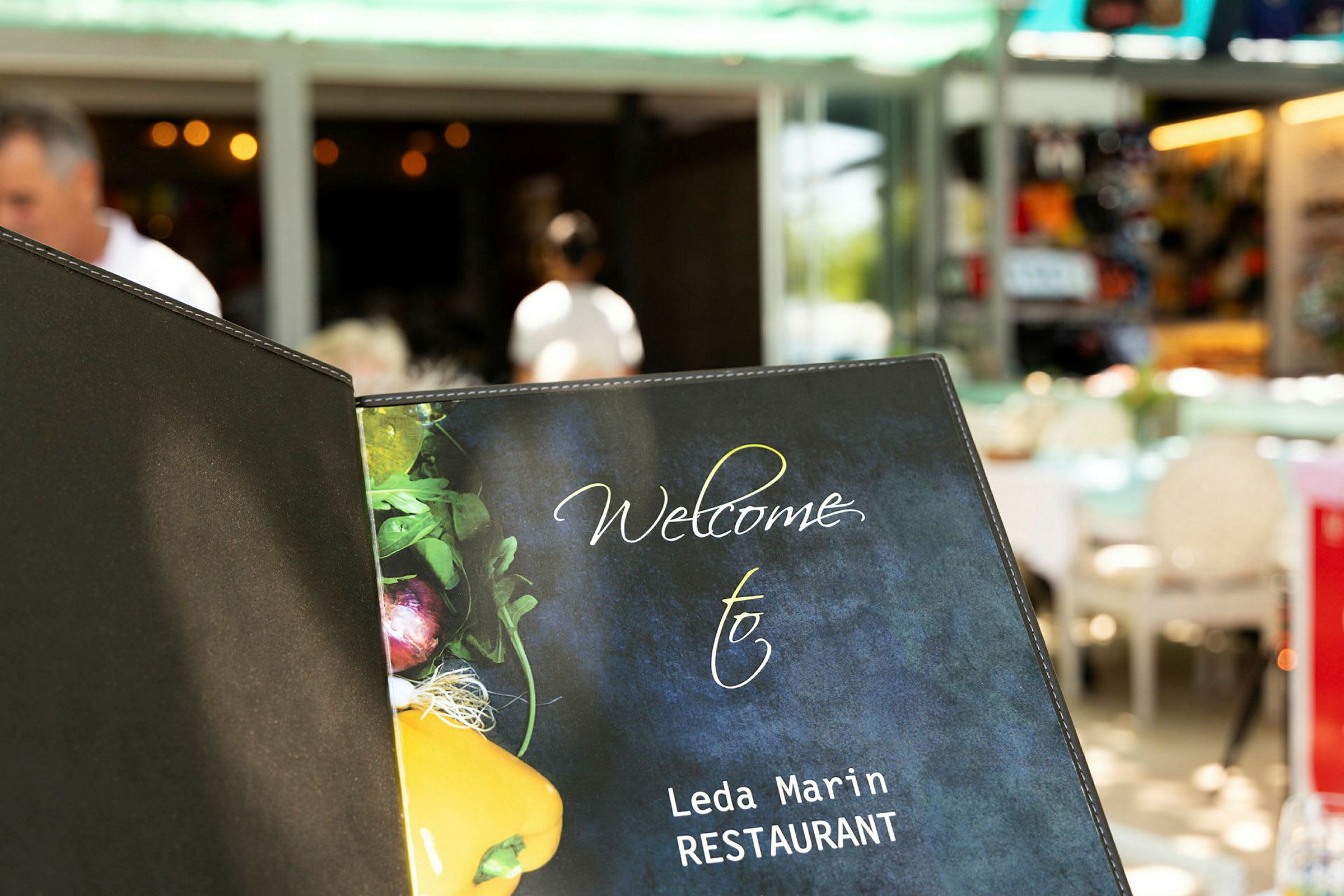 En restaurantmenuforside med teksten 'Welcome to Leda Marin RESTAURANT' og et billede af grøntsager på forsiden. (AI-genereret tekst)