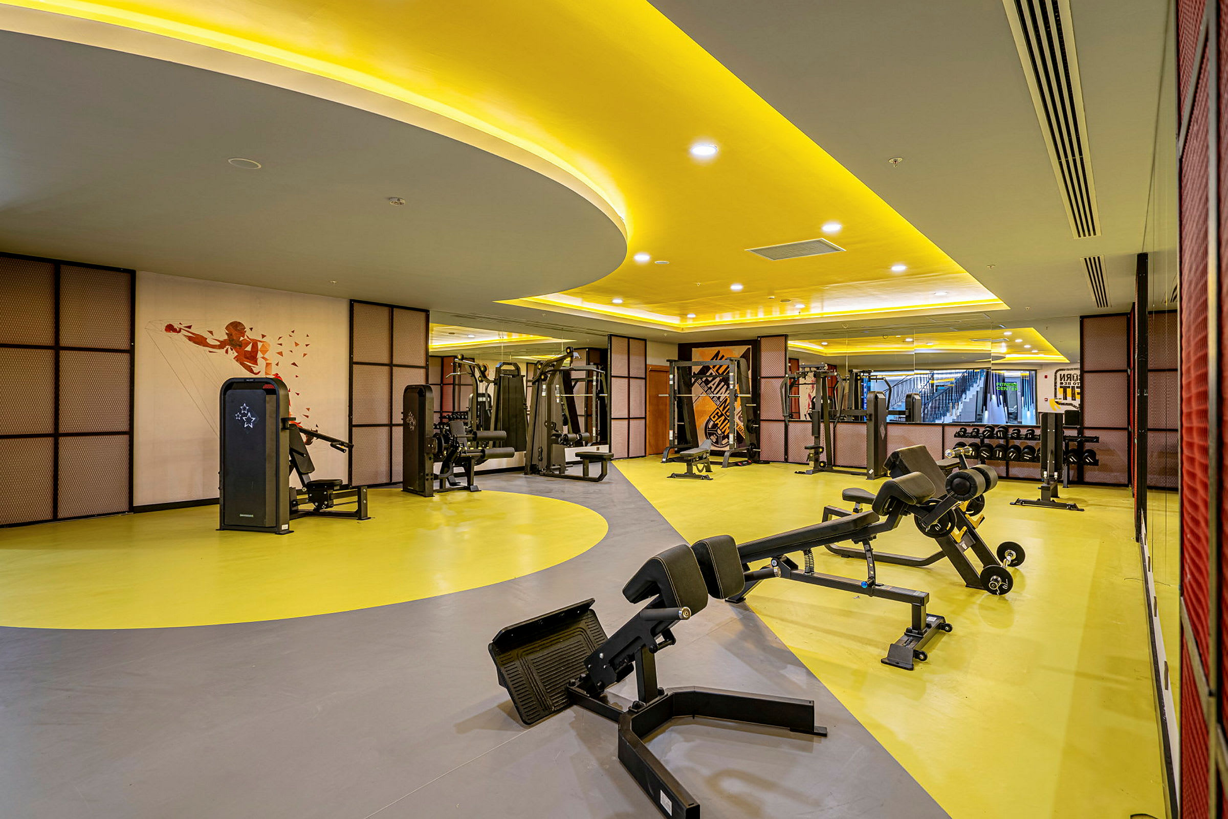 Et fitnesscenter med træningsmaskiner og bænke på et gulgråt gulv. (AI-genereret tekst)