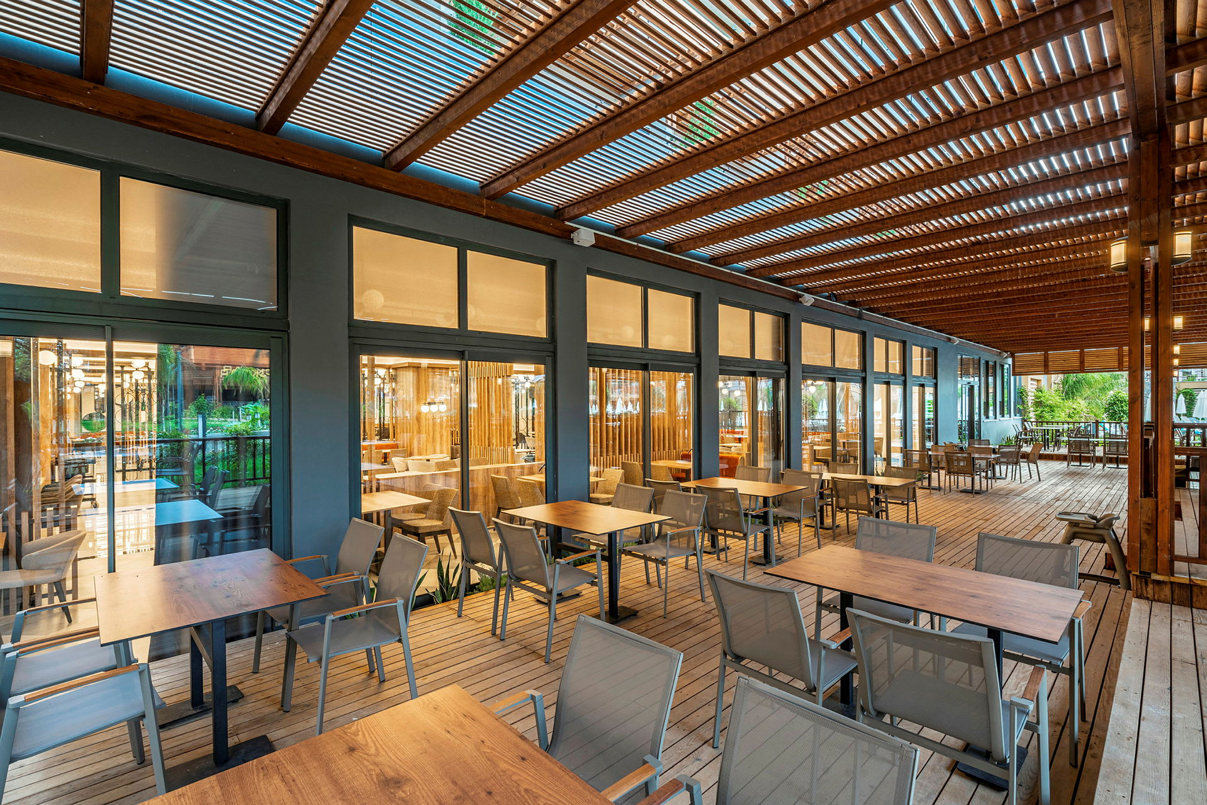 En udendørs terrasse med borde og stole, omgivet af trækonstruktion og glasvægge der vender mod en indendørs restaurant. (AI-genereret tekst)