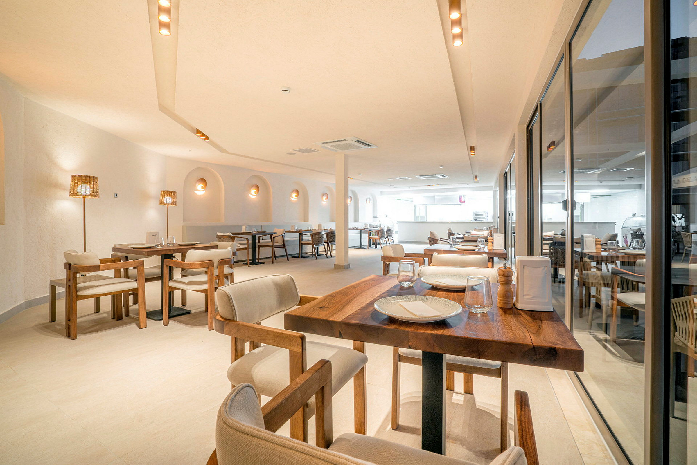 En lys og stilren restaurant med træborde, komfortable stole og dekorative lamper. Vinduer dækker den ene side af rummet. (AI-genereret tekst)