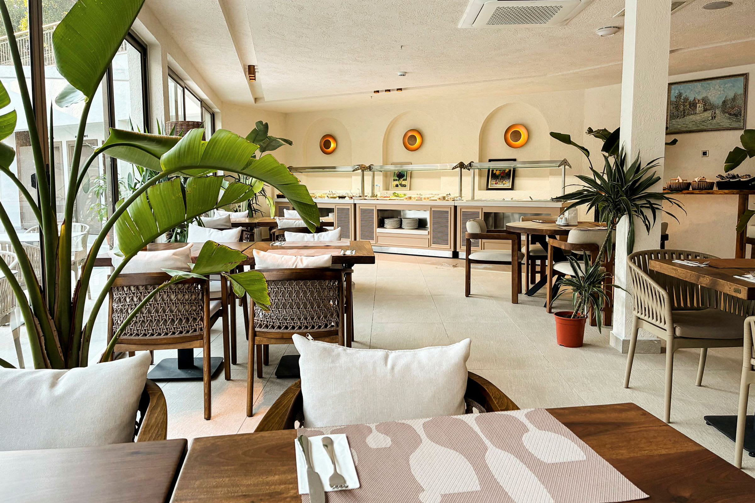 En lys og indbydende restaurant med møbler af træ, grøn planteindretning og et buffetområde i baggrunden. (AI-genereret tekst)