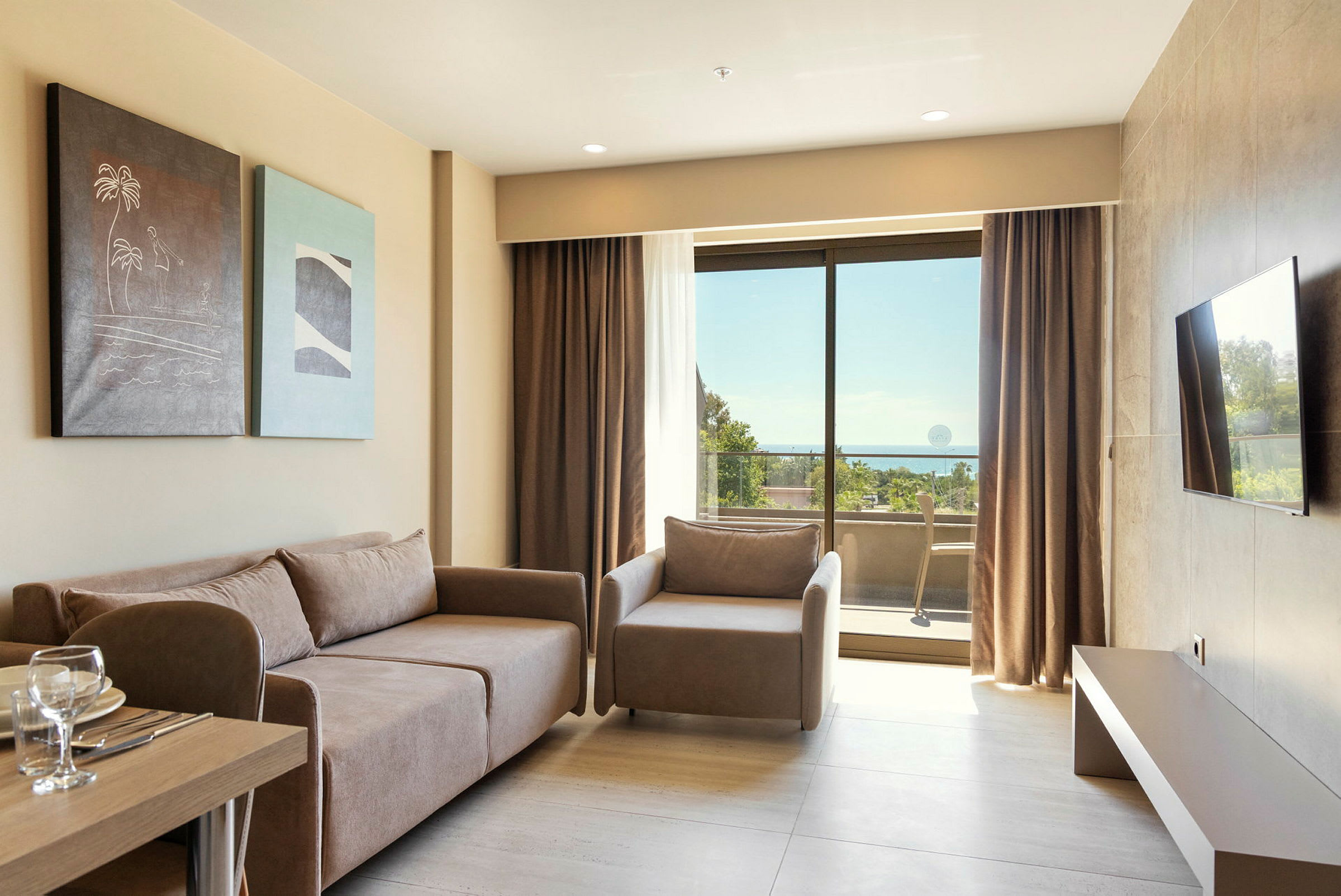 Et lyst hotelværelse med en sofa, lænestol, malerier på væggen, et bord med glas og tallerkener samt en balkon med udsigt til havet gennem store glasvinduer. (AI-genereret tekst)