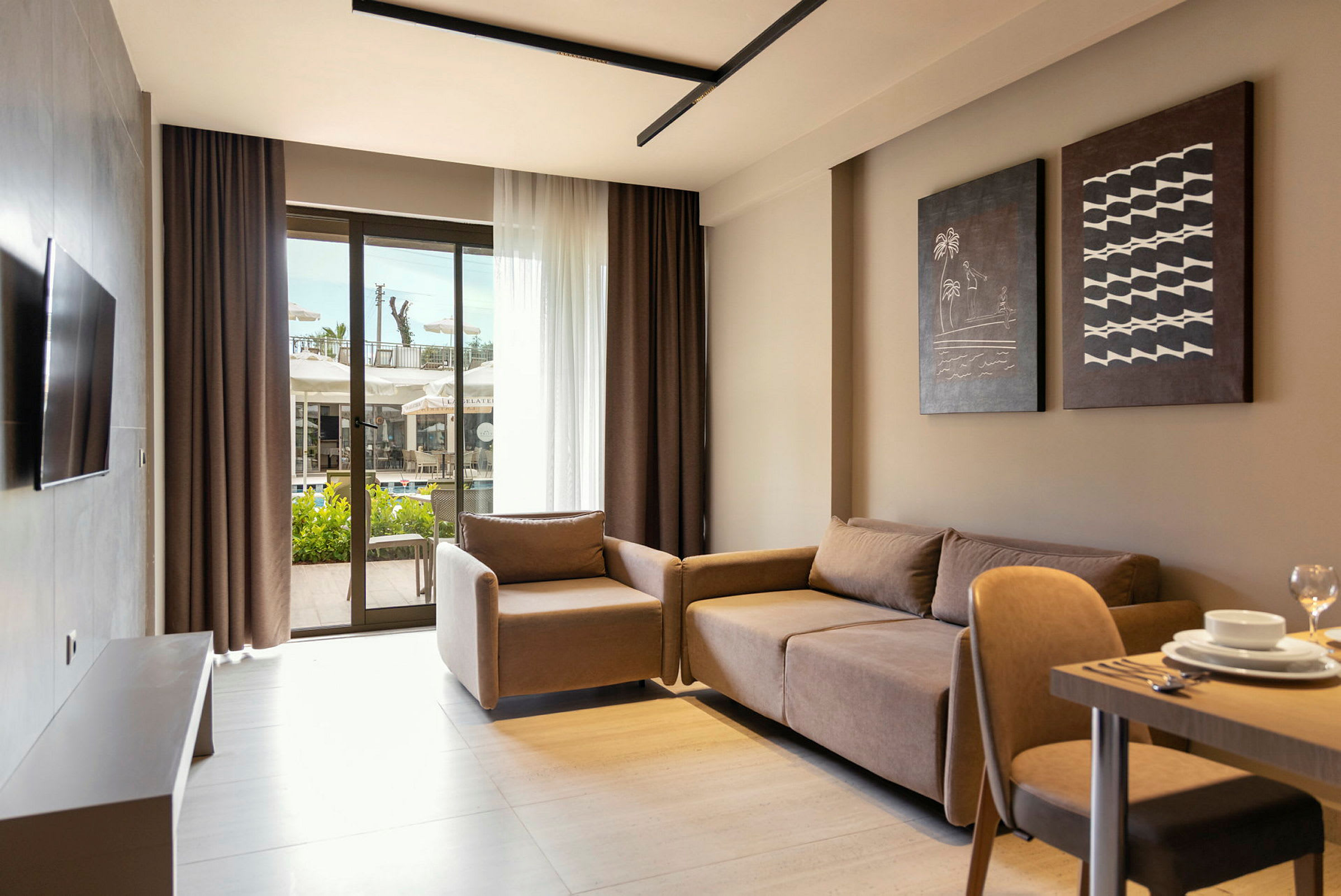 Et moderne hotelværelse med sofa, lænestol og bord set indefra. Store glasdøre i baggrunden viser udsigten til en terrasse og swimmingpool. (AI-genereret tekst)