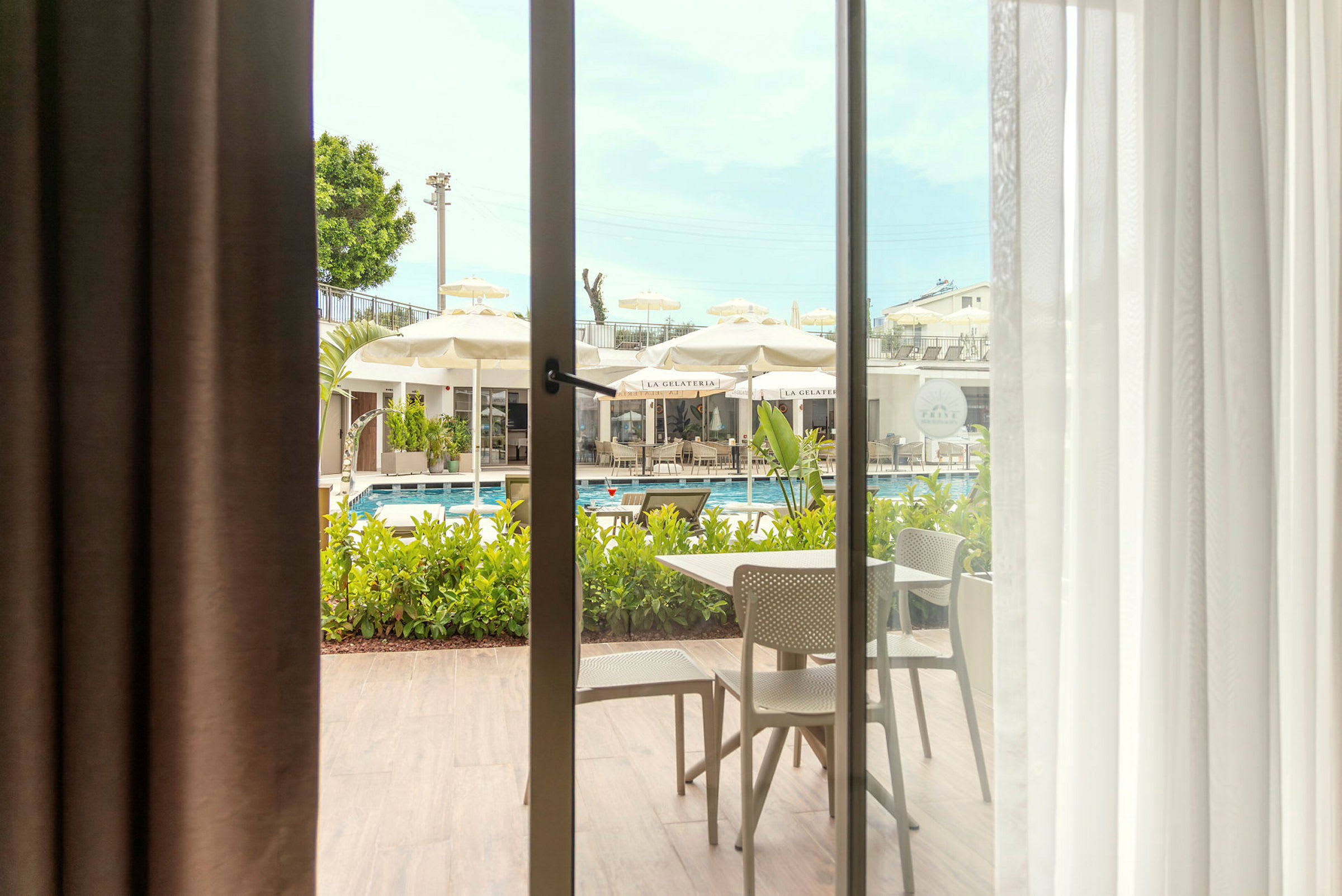 Udsigt gennem en glasdør mod en terrasse med siddemøbler og en pool omgivet af parasoller. (AI-genereret tekst)