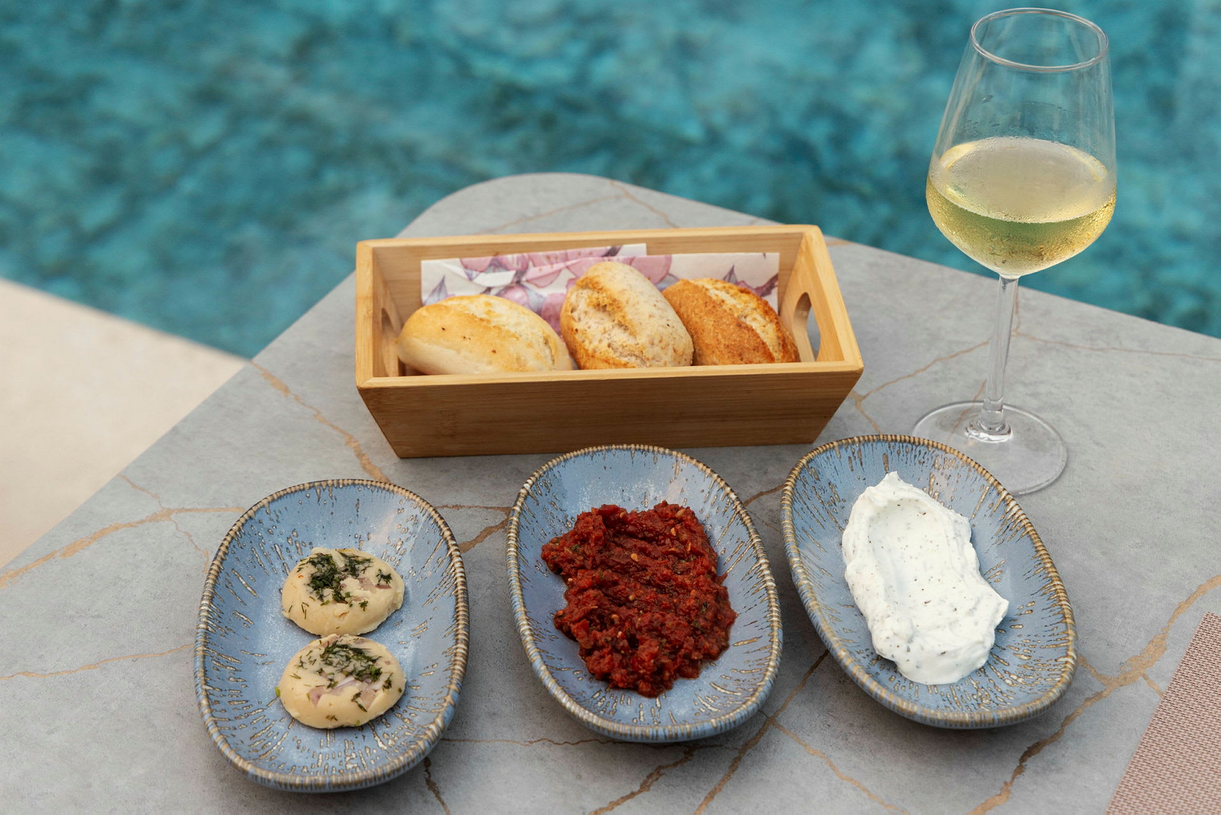En trækurv med brød, tre keramikfade med tilbehør og et glas hvidvin stående ved en pool. (AI-genereret tekst)