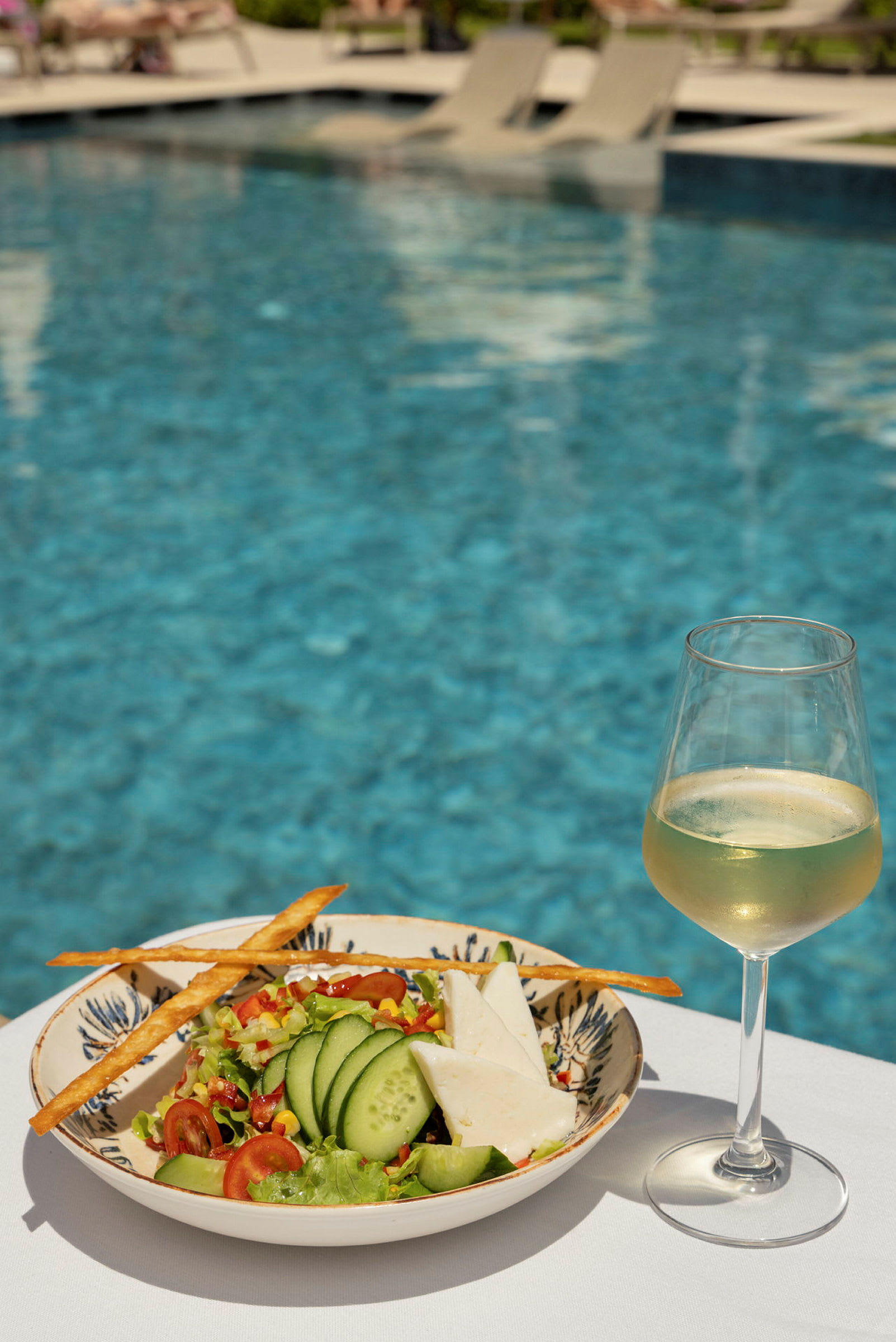 En salat med agurk og ost samt et glas hvidvin står på et bord med en pool i baggrunden. (AI-genereret tekst)