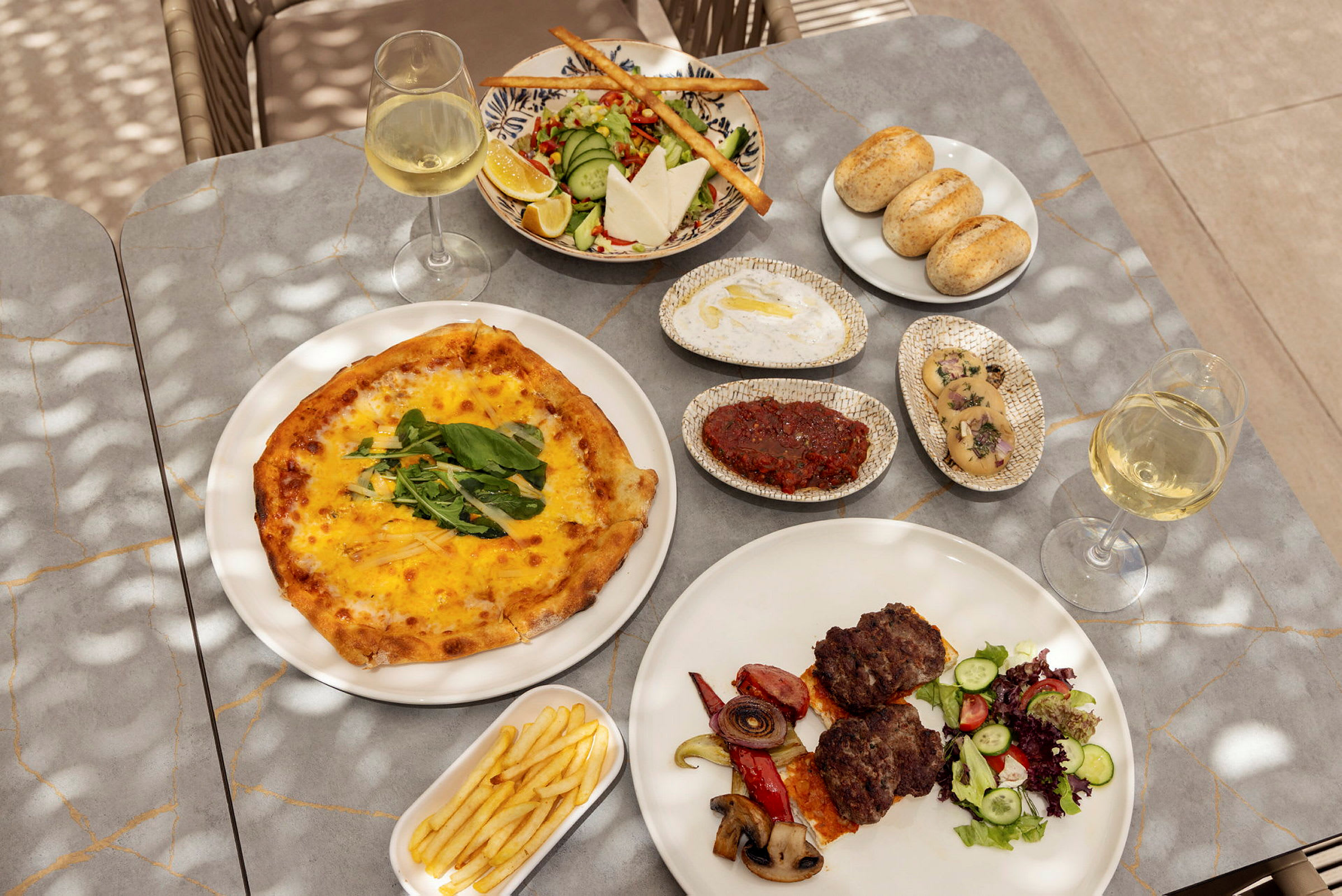 Et bord med forskellige retter, herunder pizza, salat, brød, kød med grøntsager, pommes frites samt glas med hvidvin. (AI-genereret tekst)