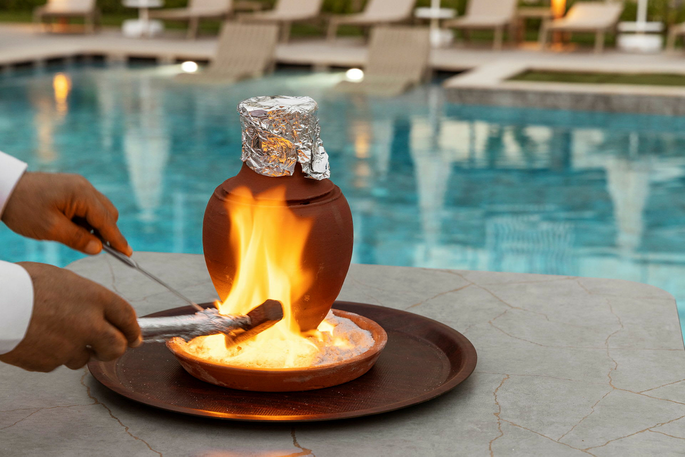 Et bord med en lerkrukke dækket af folie står på en bakke, med en flamme tændt nedenunder. I baggrunden ses en pool og solsenge. (AI-genereret tekst)
