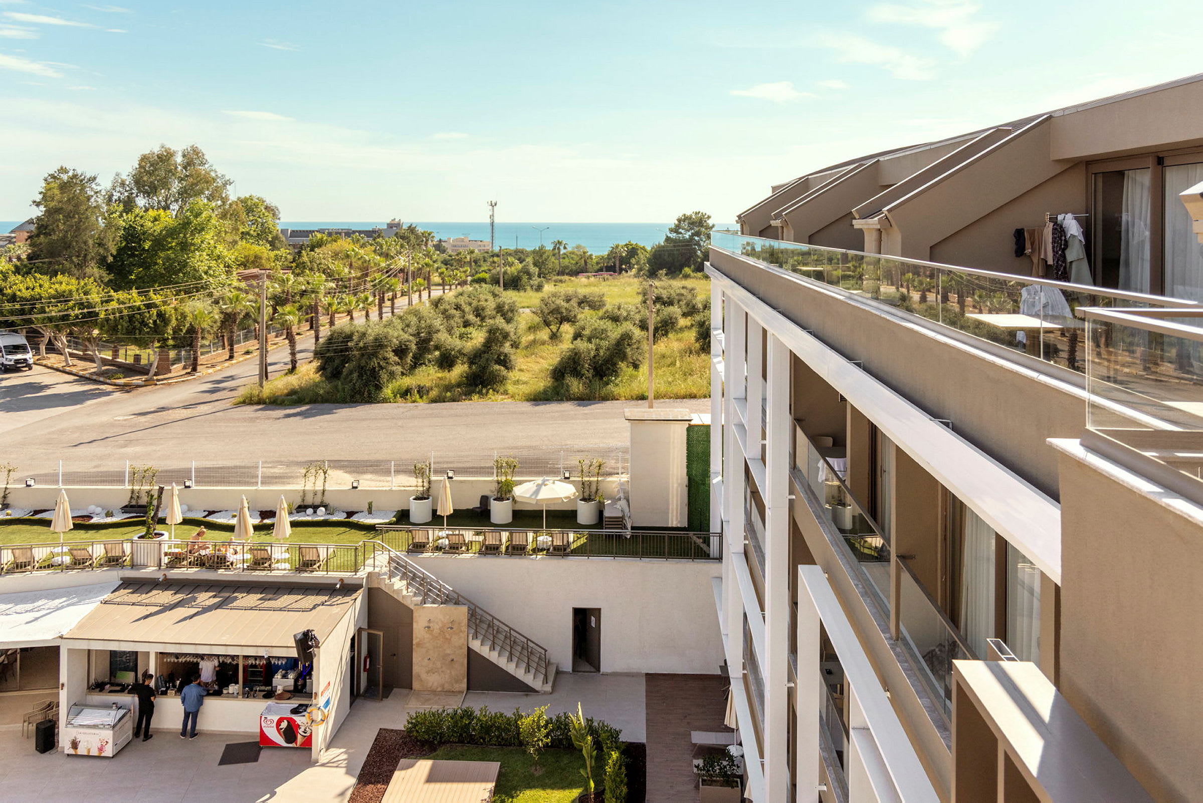 Et hotel med moderne arkitektur og altaner, beliggende ved siden af et trælæsket område og en vej. I baggrunden kan havet ses. (AI-genereret tekst)
