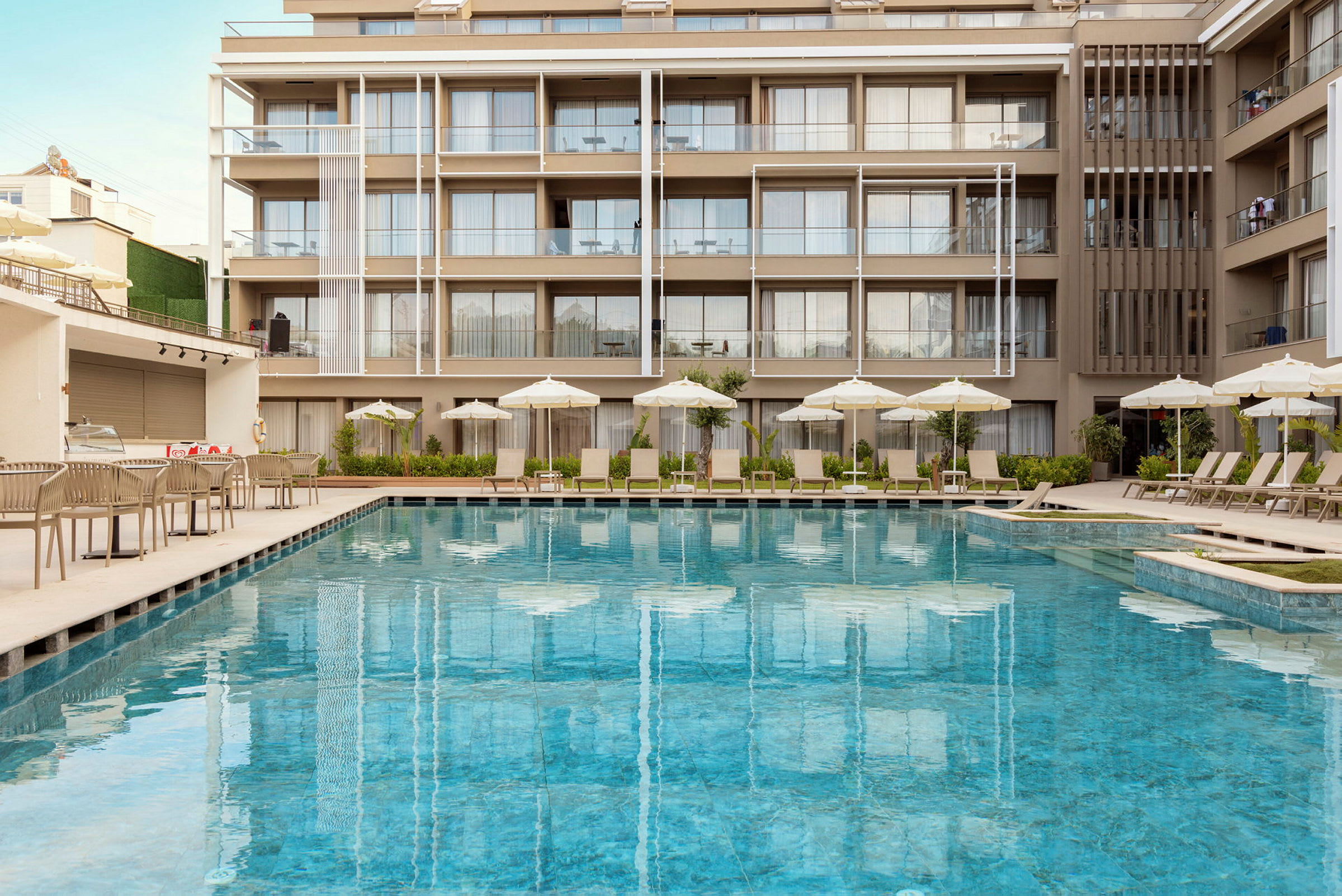 Hotel med en udendørs pool omgivet af liggestole og parasoller. Baggrunden viser altaner og moderne arkitektur. (AI-genereret tekst)