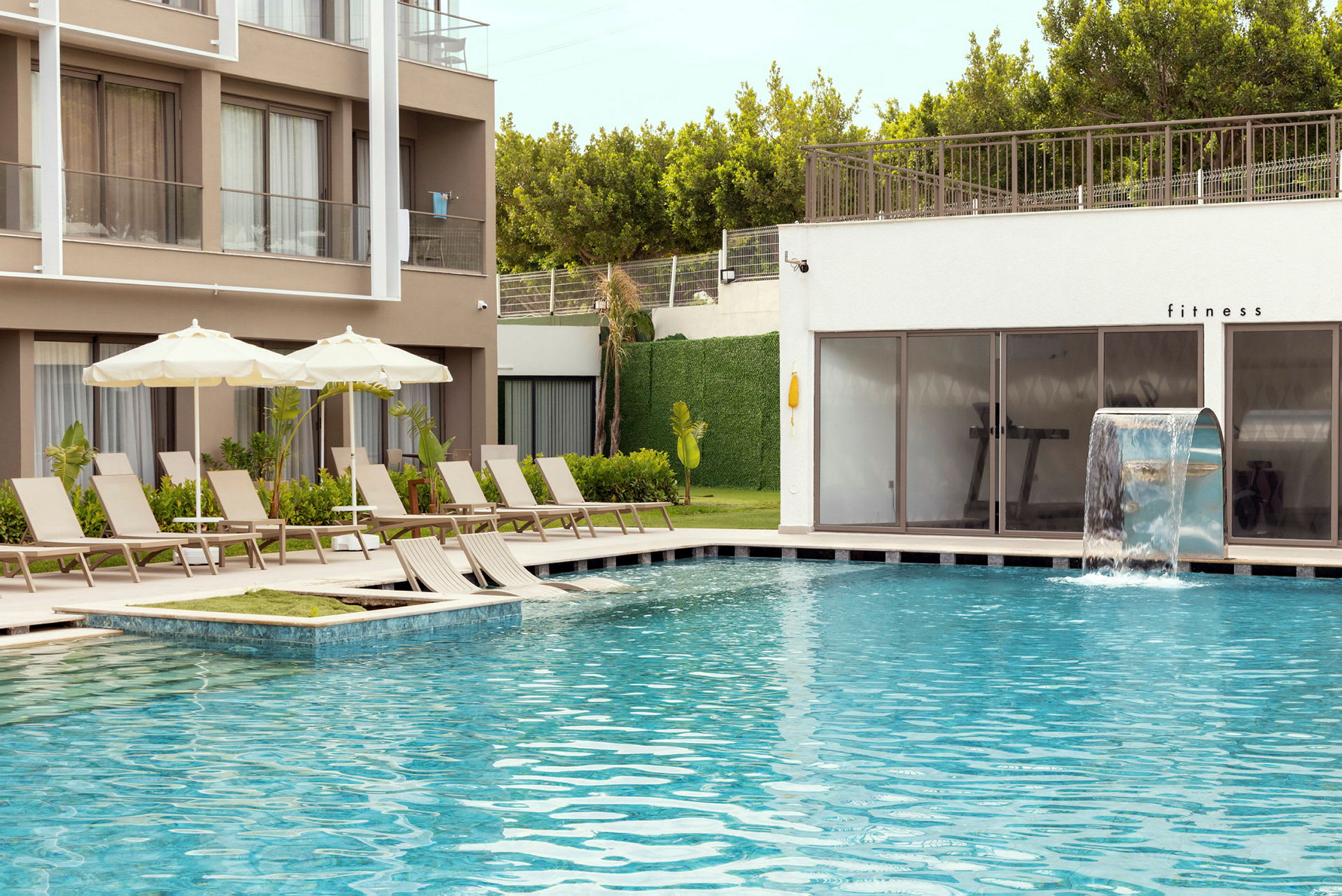 Udendørs swimmingpool med liggestole og parasoller ved et hotel eller resort. En vandfaldsfunktion er til stede i poolen, og et fitnessrum kan ses i baggrunden. (AI-genereret tekst)