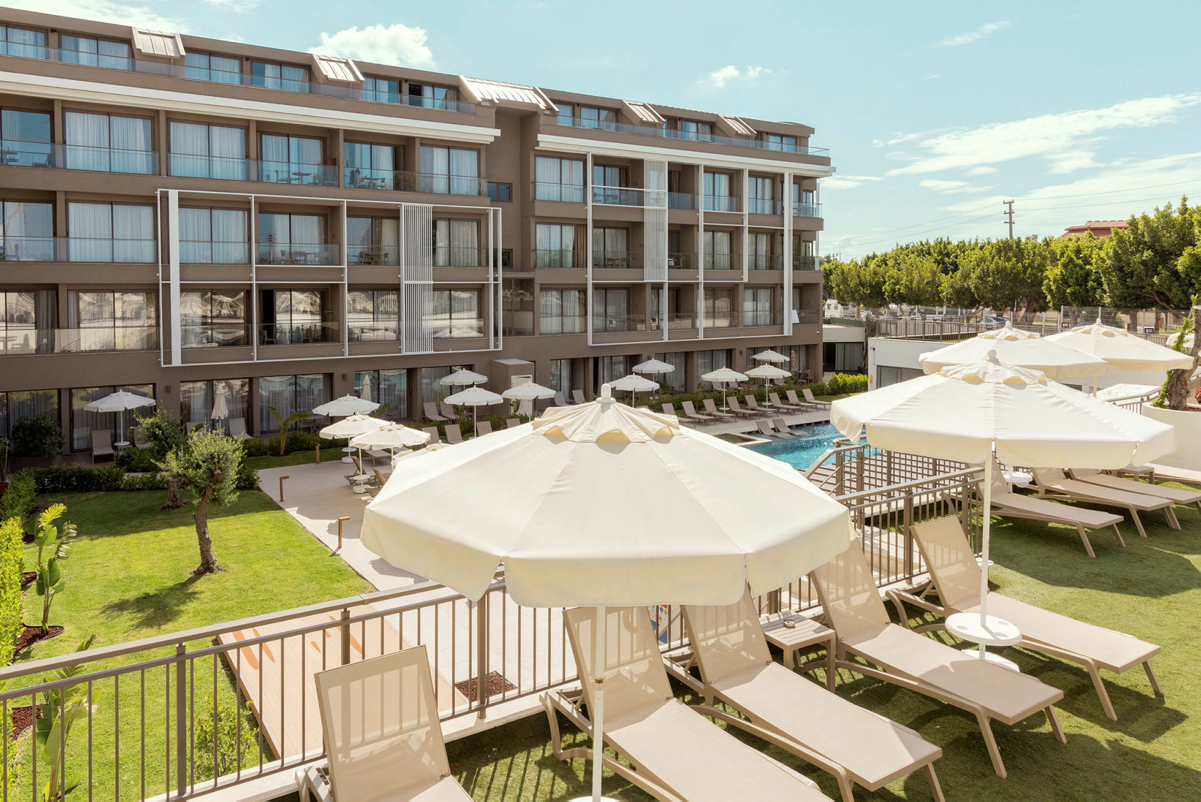 En moderne hotelbygning med flere etager. Foran bygningen er der en græsplæne, liggestole og parasoller ved en swimmingpool. (AI-genereret tekst)