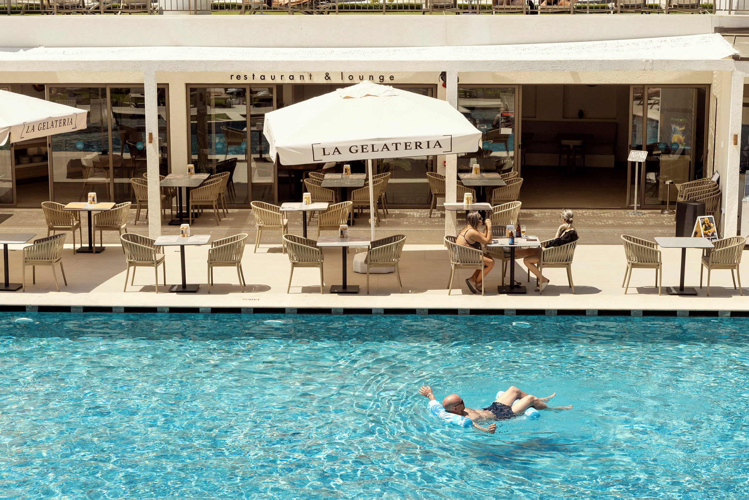 En person flyder i en pool foran en restaurantterrasse med parasoller og siddepladser. (AI-genereret tekst)