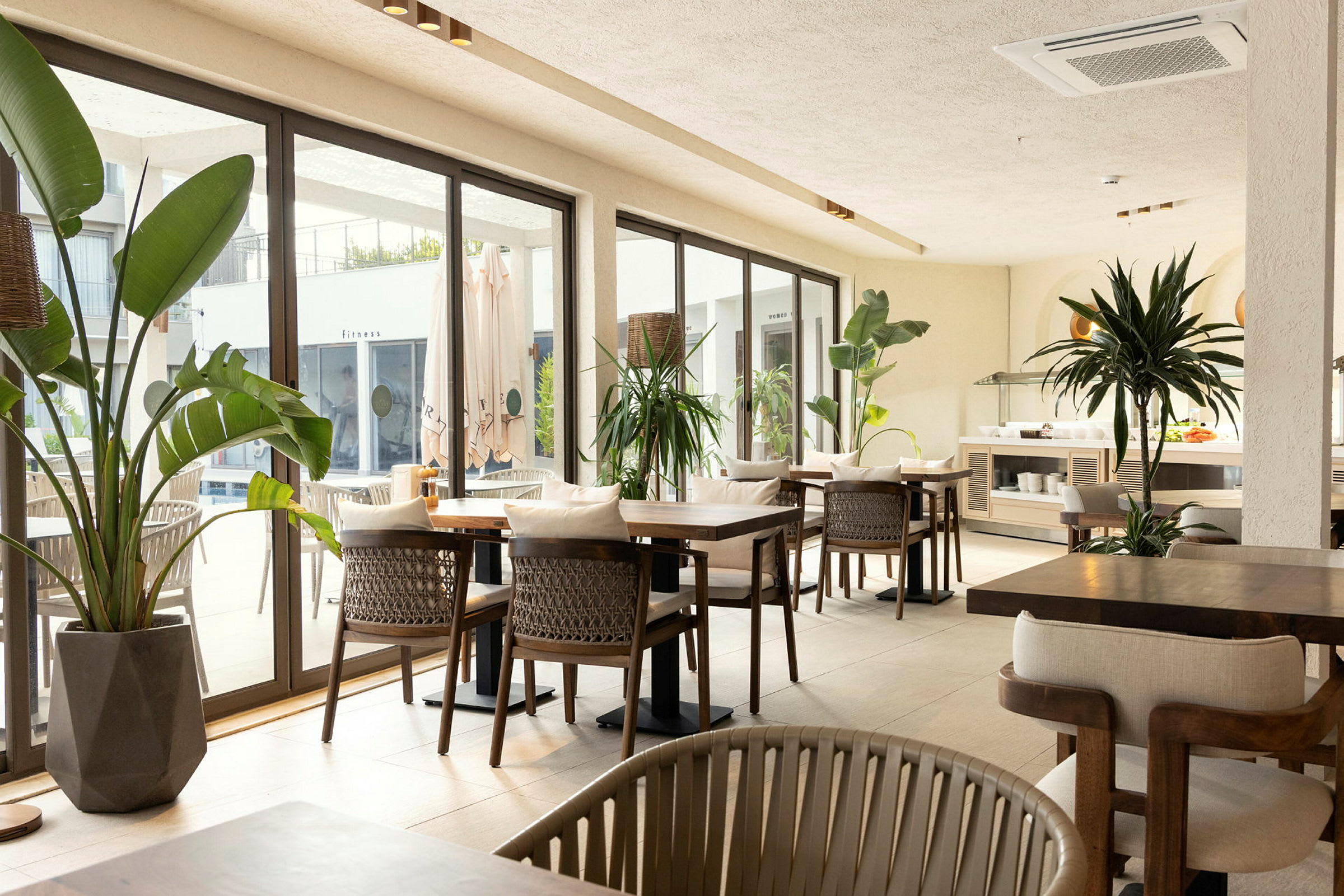 Restaurantområde med borde og stole, omgivet af store vinduer og grønne planter. (AI-genereret tekst)