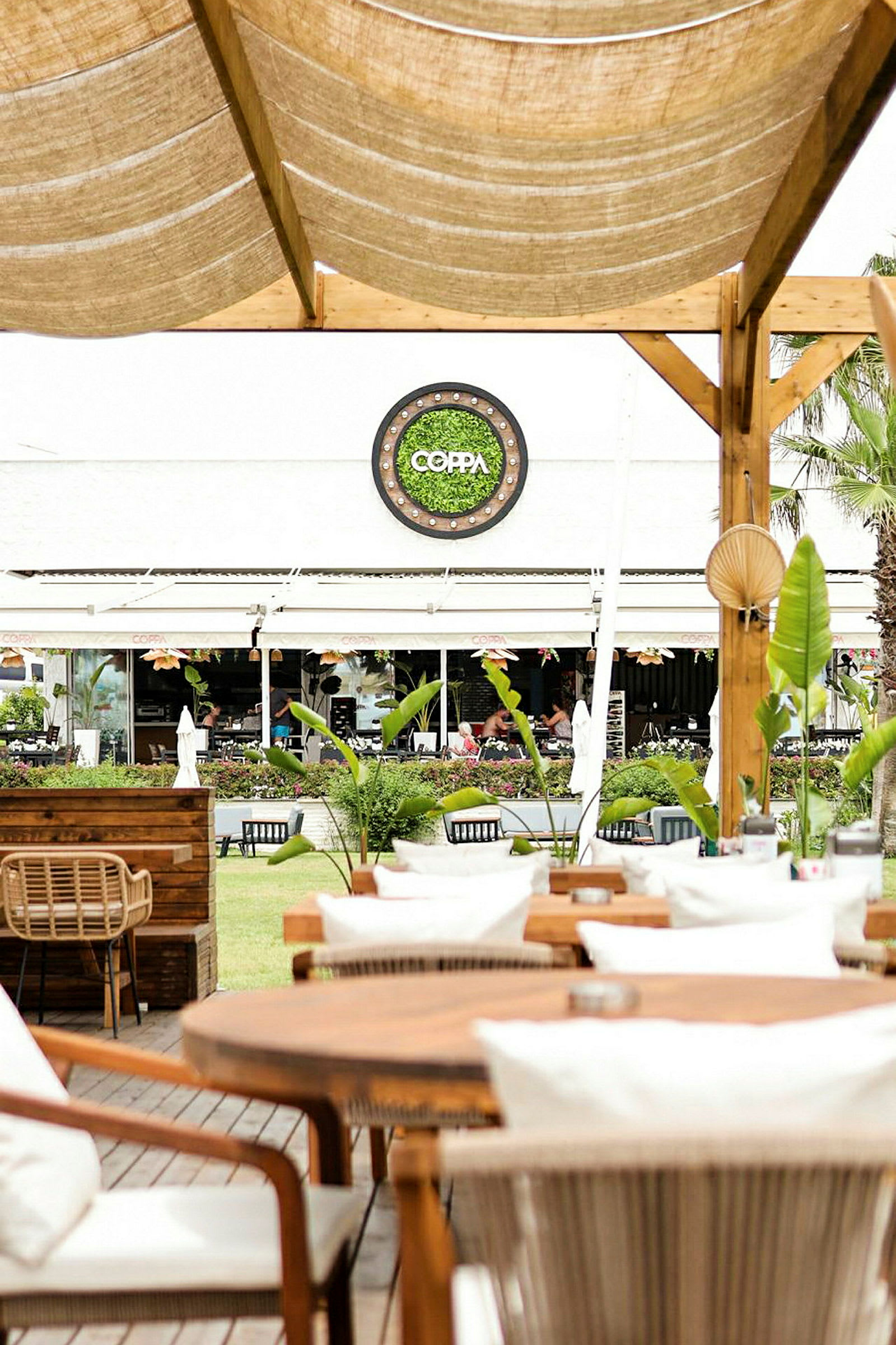 Udendørs servering med træborde og stole, have og restauranten Coppa i baggrunden. (AI-genereret tekst)