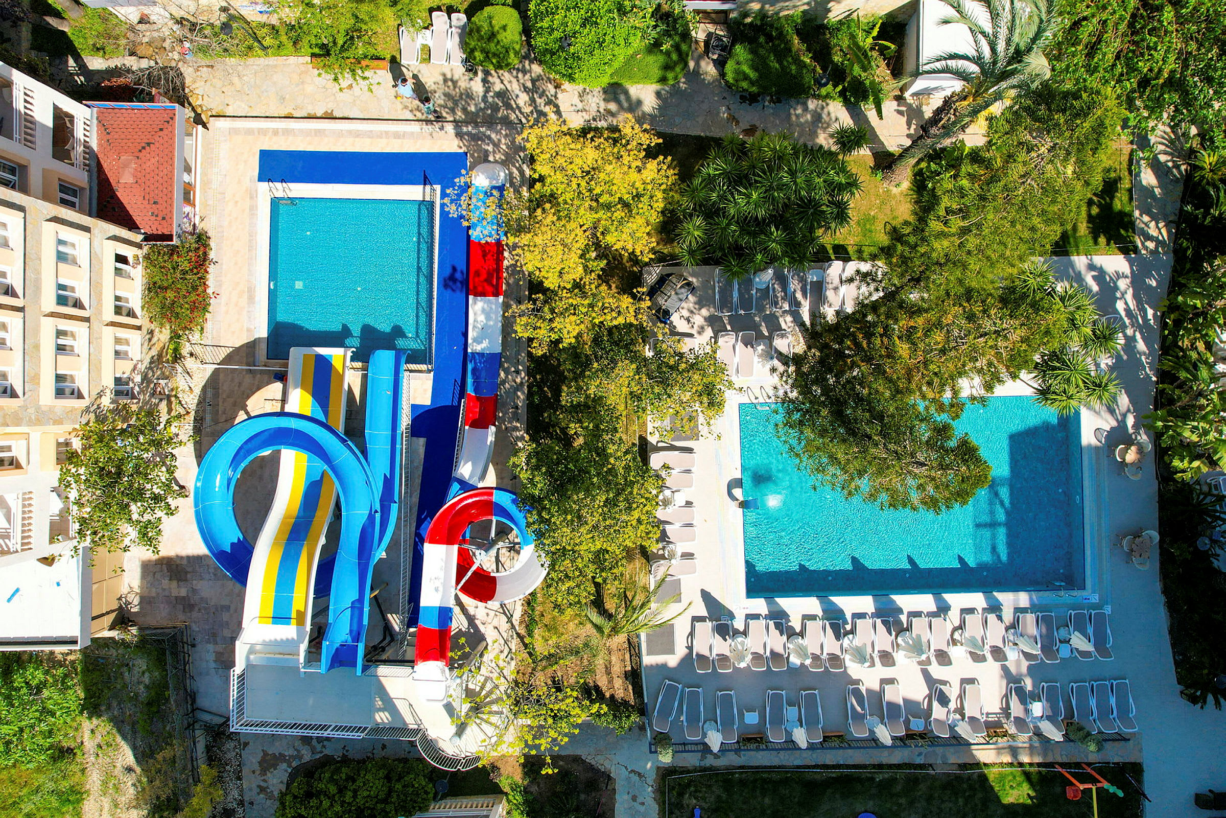 To pools, hvoraf den ene har vandrutsjebaner, omringet af liggestole og grønne områder. (AI-genereret tekst)