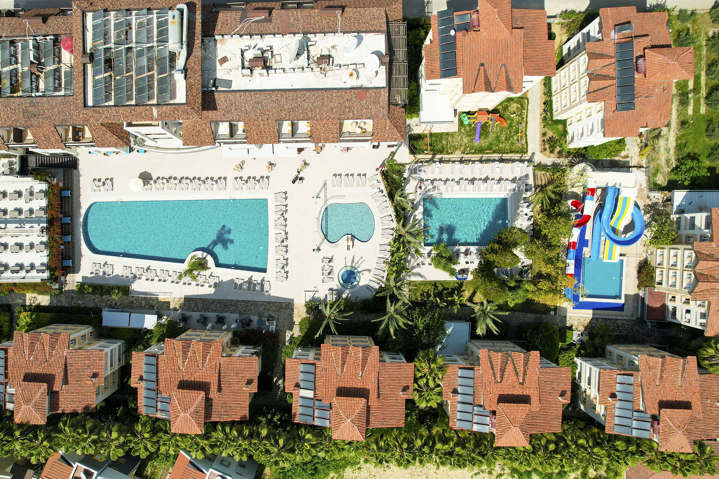 Luftfoto af hotelkompleks med pools og vandrutsjebaner, omgivet af bygninger med røde tage. (AI-genereret tekst)