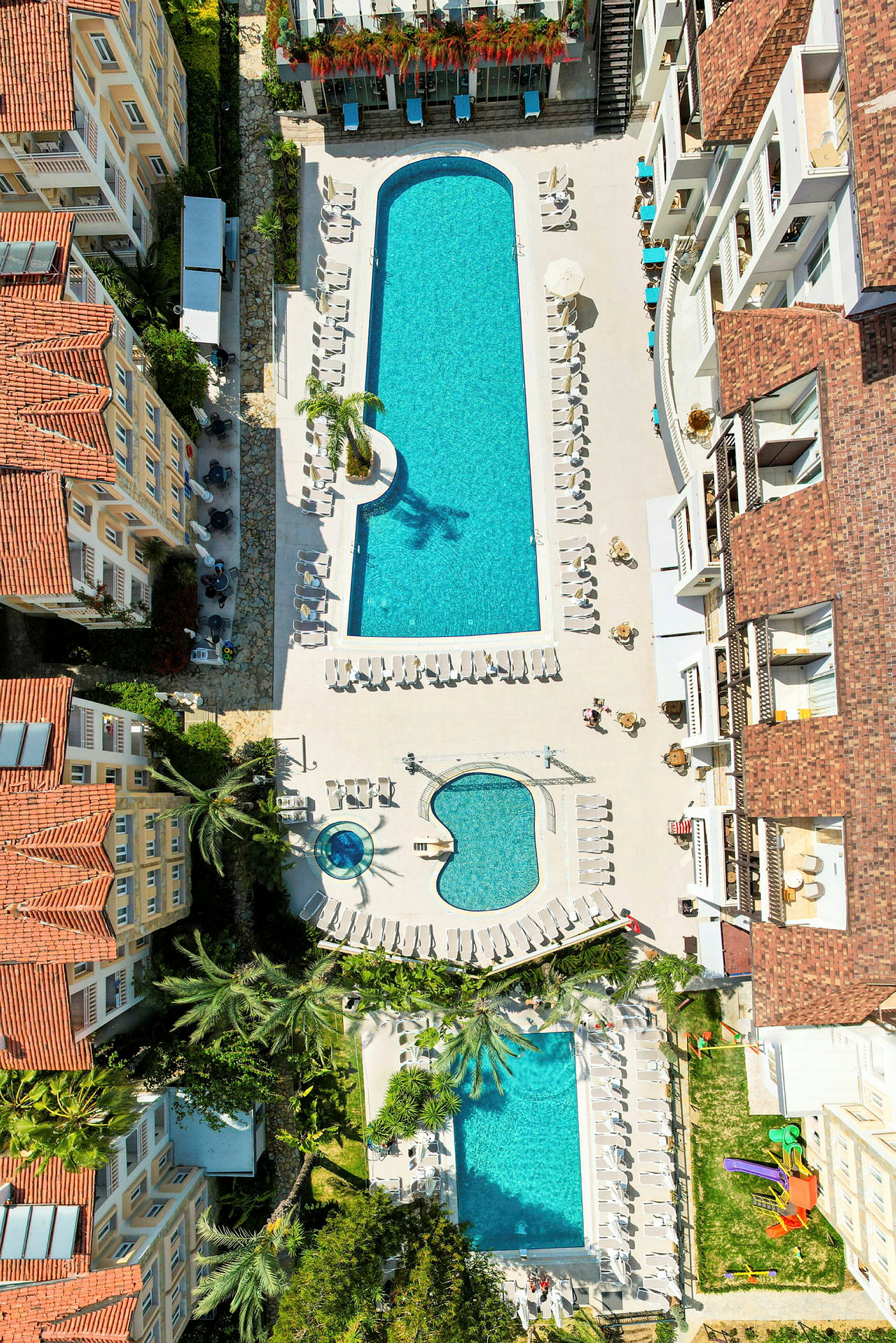Fugleperspektiv af hotelområde med flere swimmingpools og liggestole. (AI-genereret tekst)