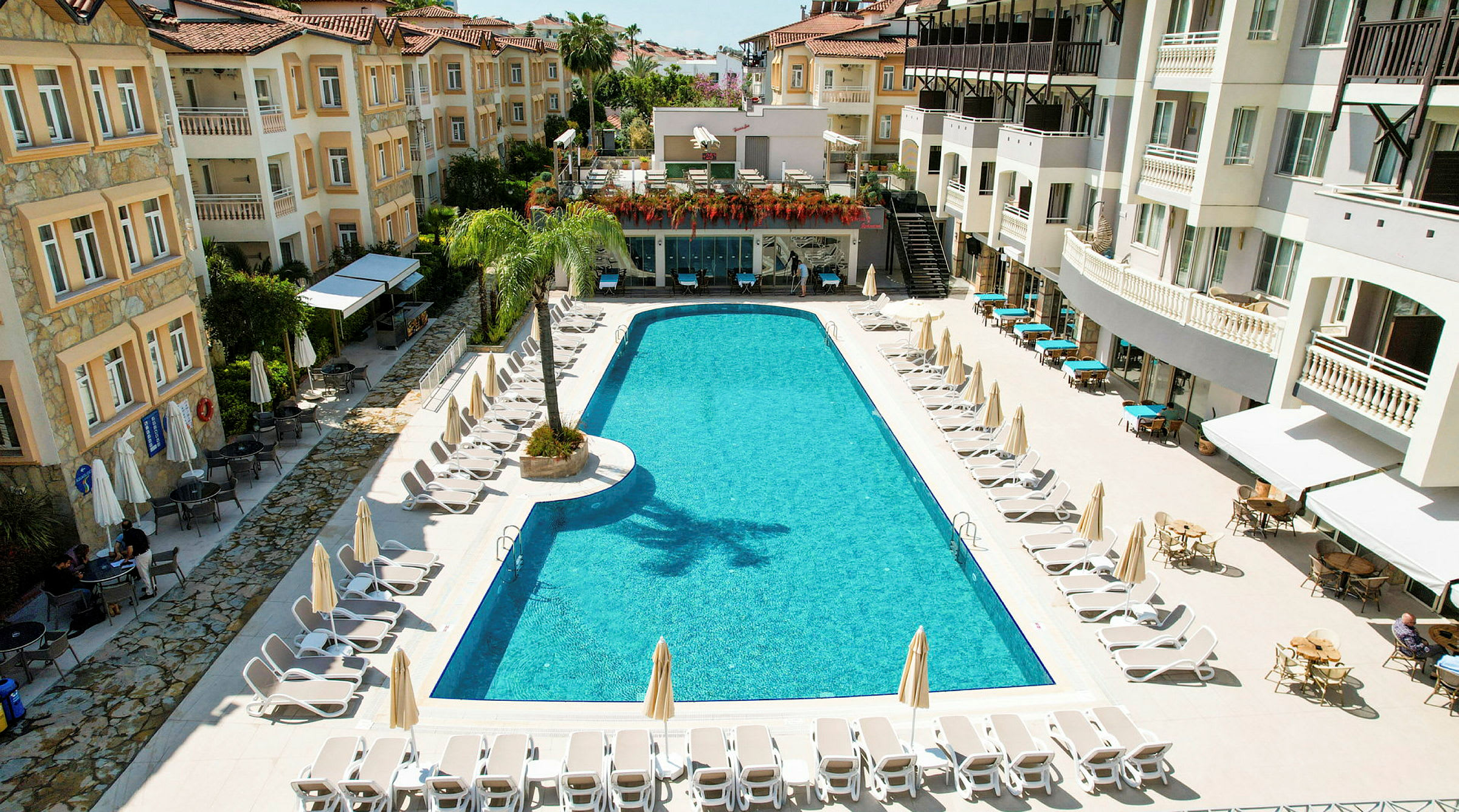 Hotelområde med en lang swimmingpool omgivet af solsenge og parasoller, omgivet af bygninger med altaner. (AI-genereret tekst)