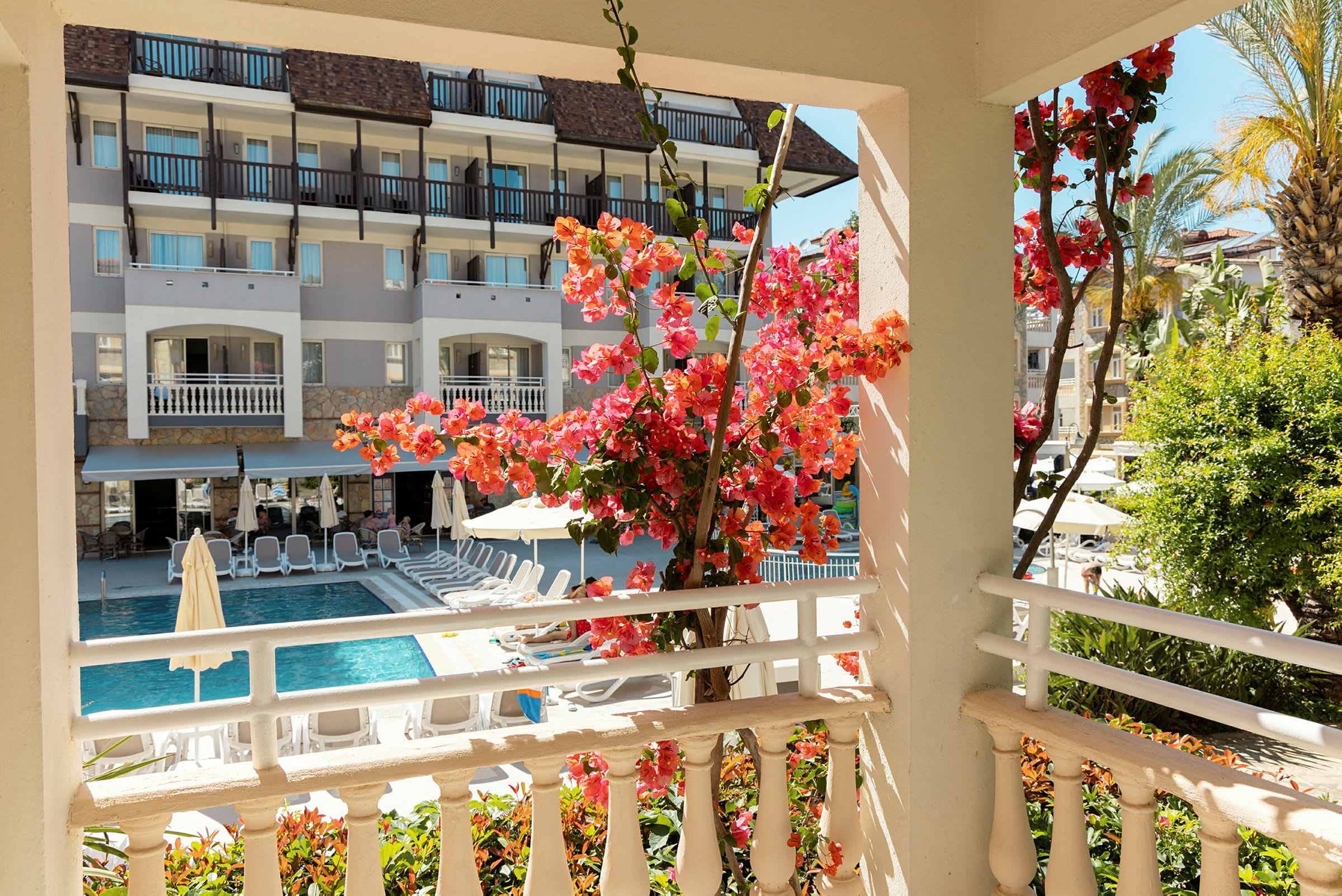Udsigt fra en terrasse mod en pool omgivet af liggestole, med blomstrende bougainvillea i forgrunden. (AI-genereret tekst)