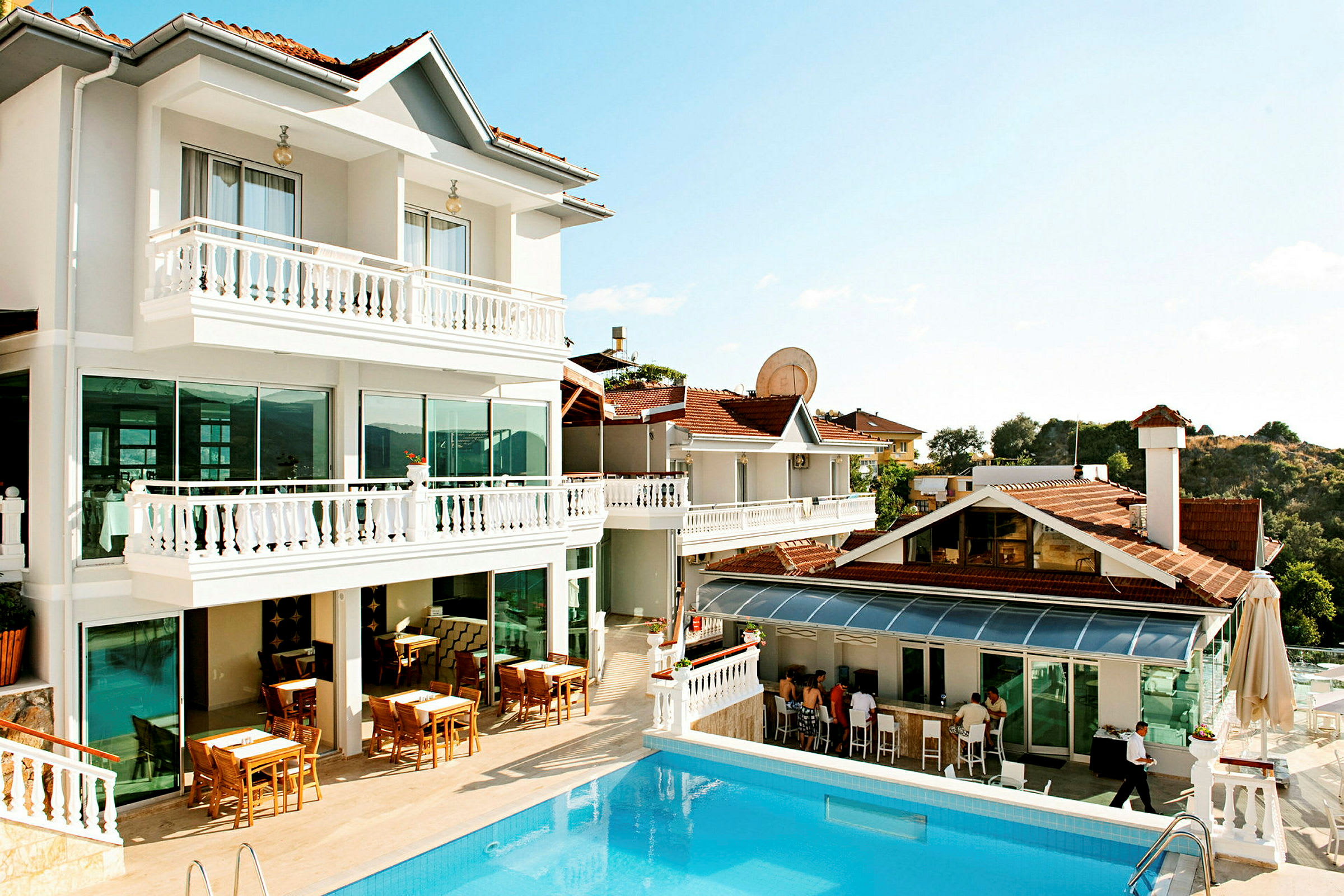 Hotelbygning med hvid facade, udendørs borde og swimmingpool. (AI-genereret tekst)