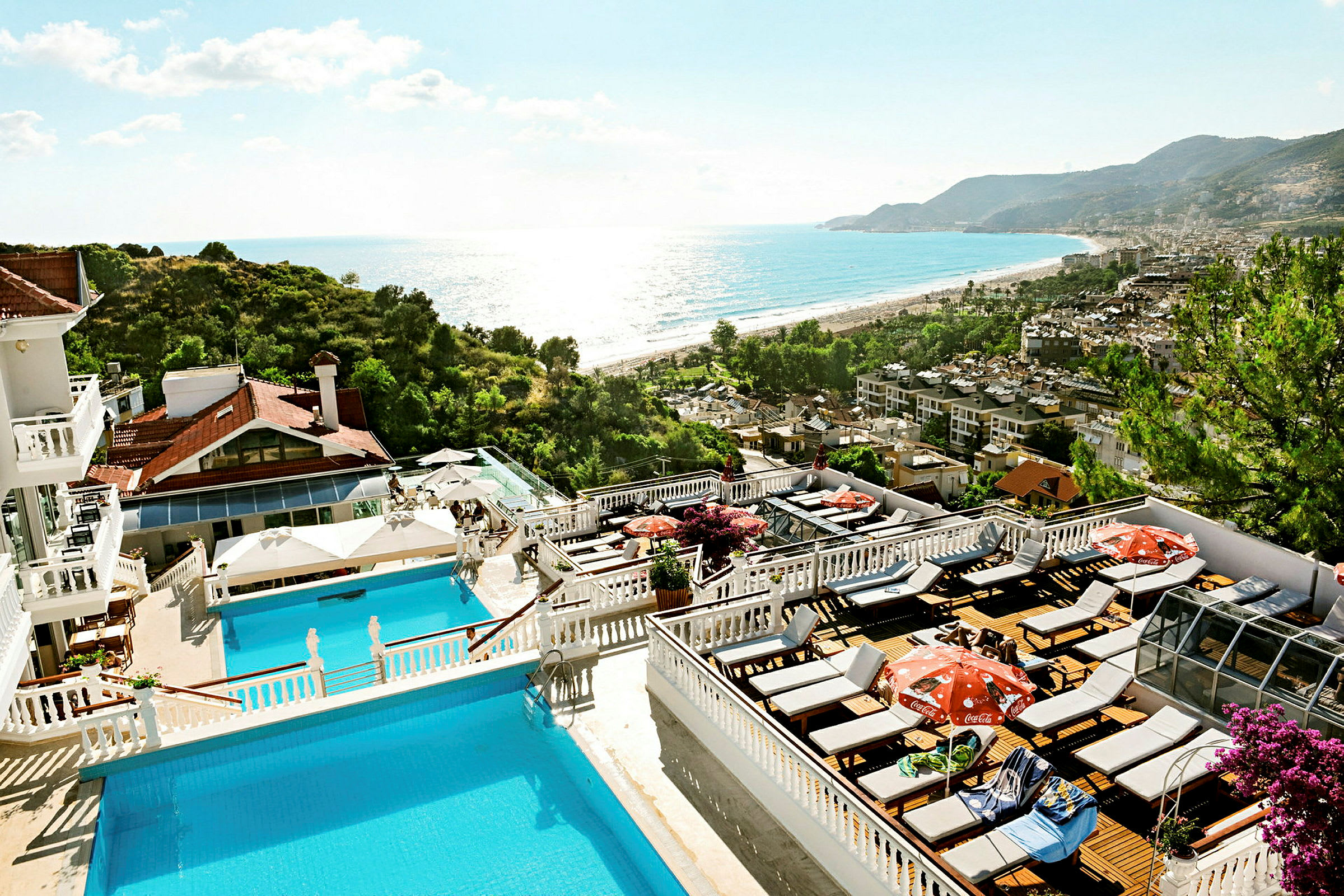Billede af et hotelområde med pools og solterrasser, omgivet af grønne områder og med udsigt over havet og en strand. (AI-genereret tekst)