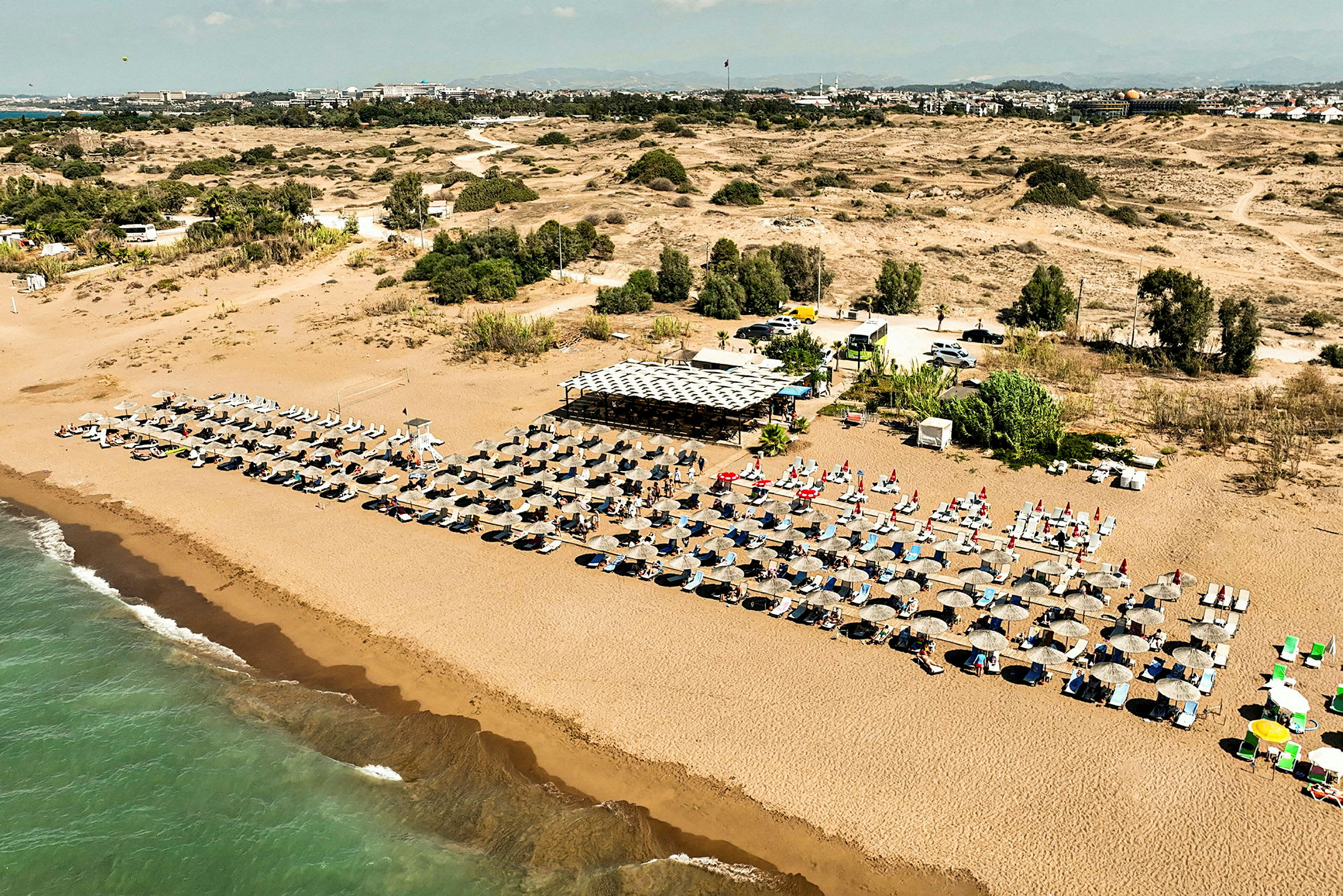 En lang sandstrand med rækker af liggestole og parasoller samt en strandcafé i baggrunden. (AI-genereret tekst)