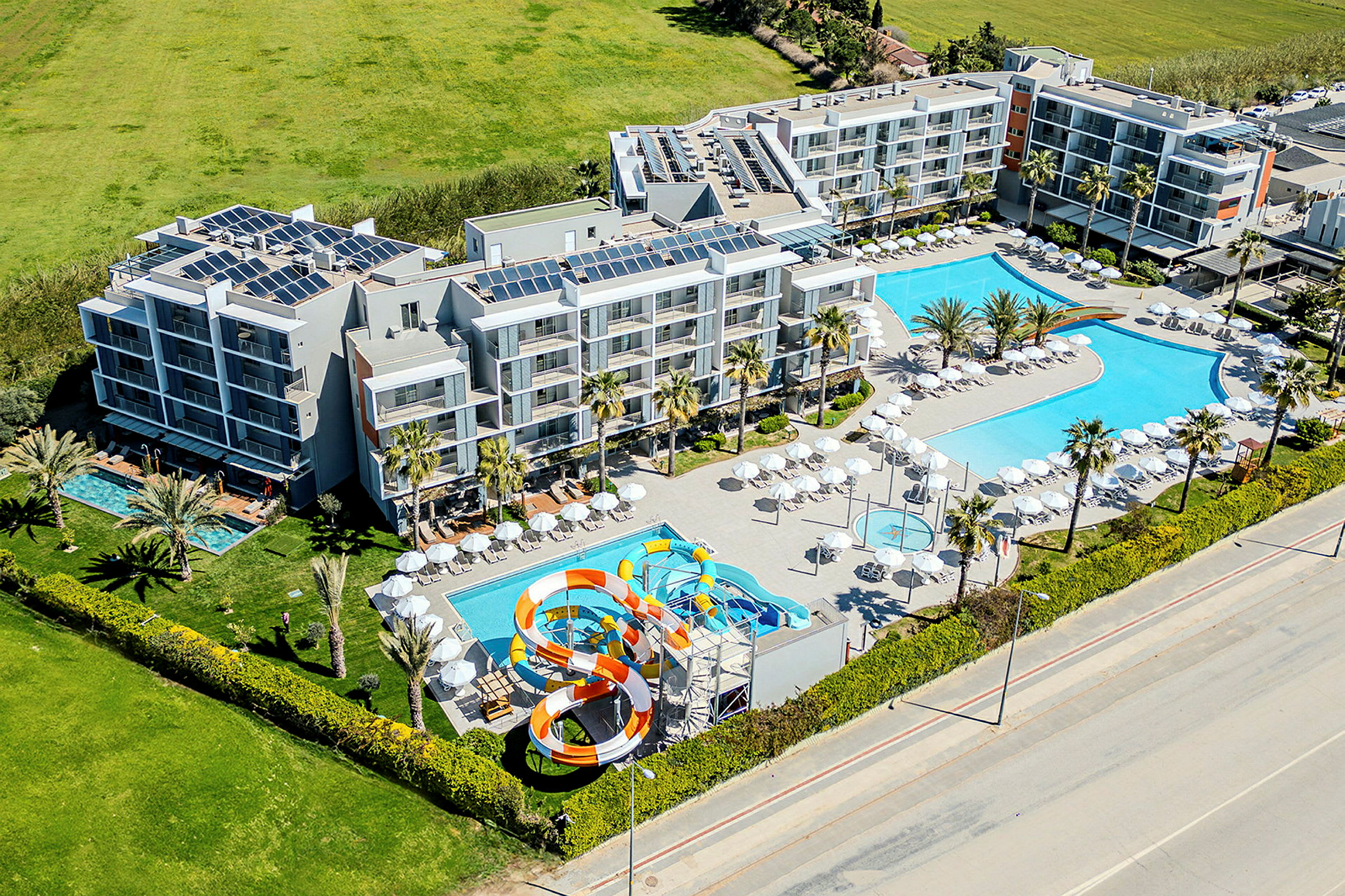 Hotelkompleks med flere bygninger, to store poolområder og en vandrutsjebane, omgivet af grønt og en vej. (AI-genereret tekst)