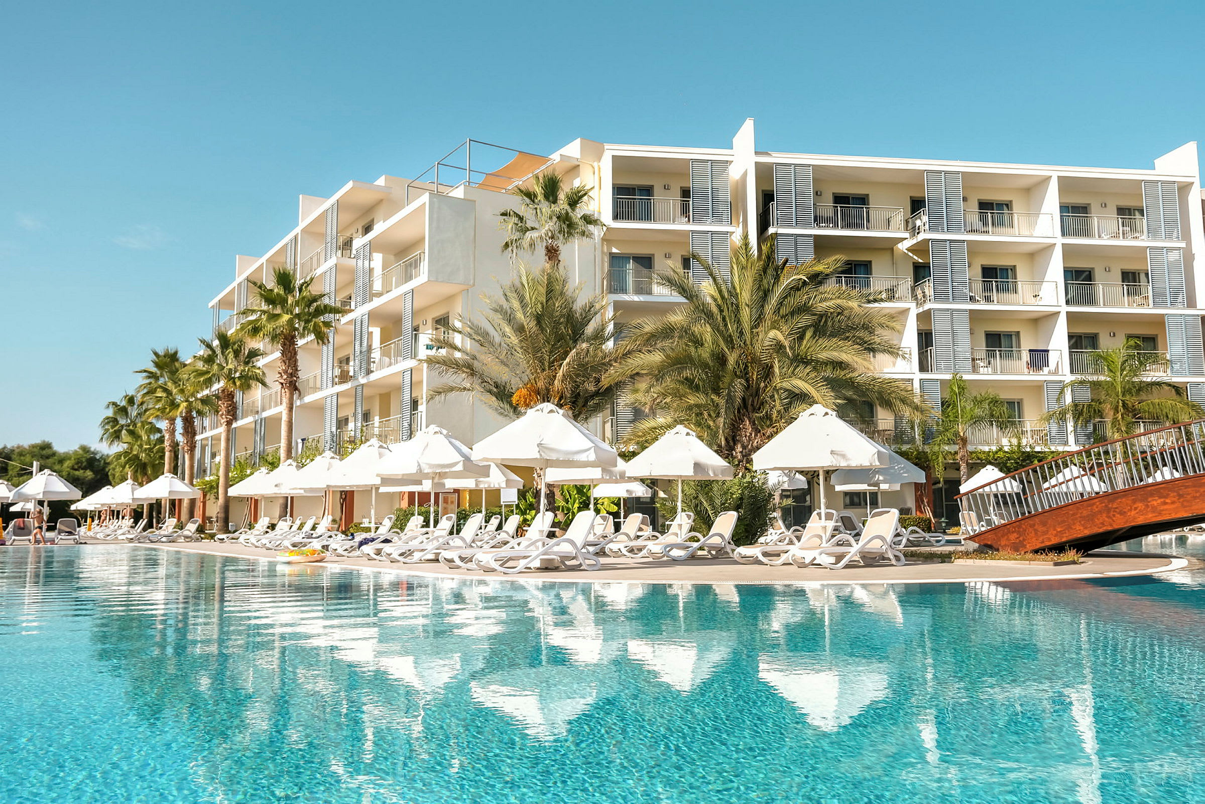 En hotelbygning ved siden af en stor pool med liggestole og parasoller. (AI-genereret tekst)