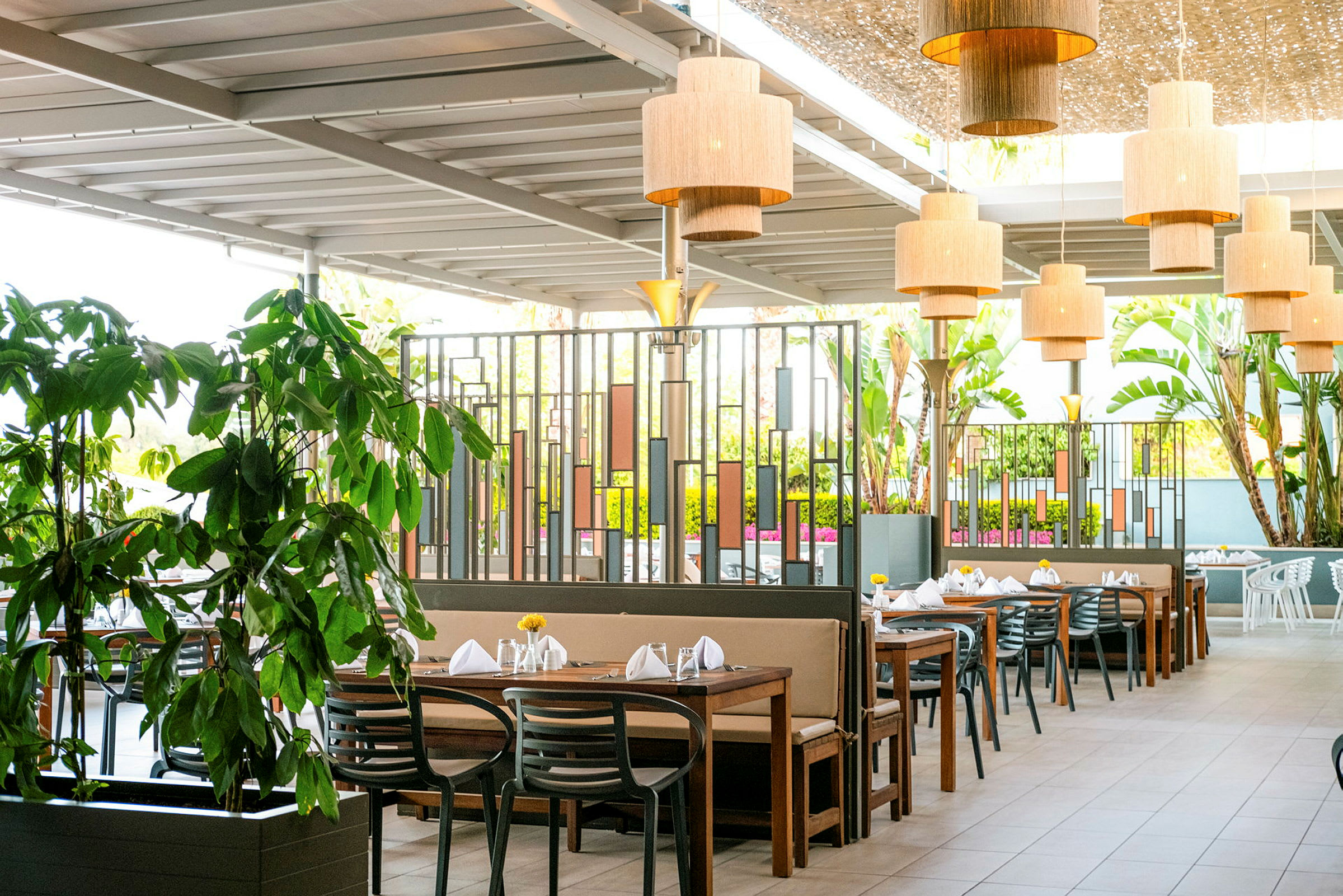 Billede af en udendørs restaurant med flere borde, stole og grønne planter. (AI-genereret tekst)